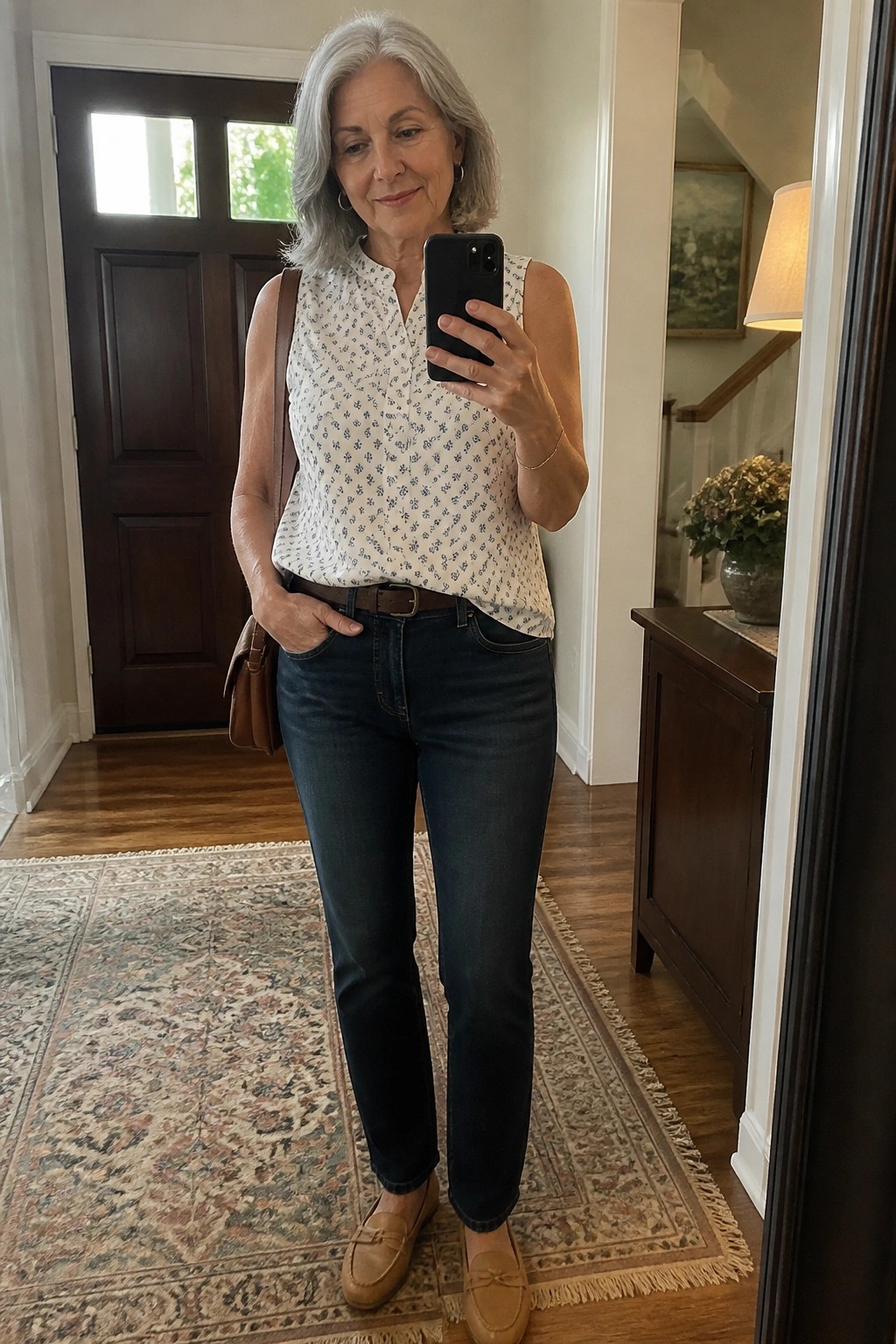 Woman over 50 wearing a sleeveless white top with small blue floral print tucked into dark blue jeans with a brown belt, paired with brown loafers and carrying a brown shoulder bag.