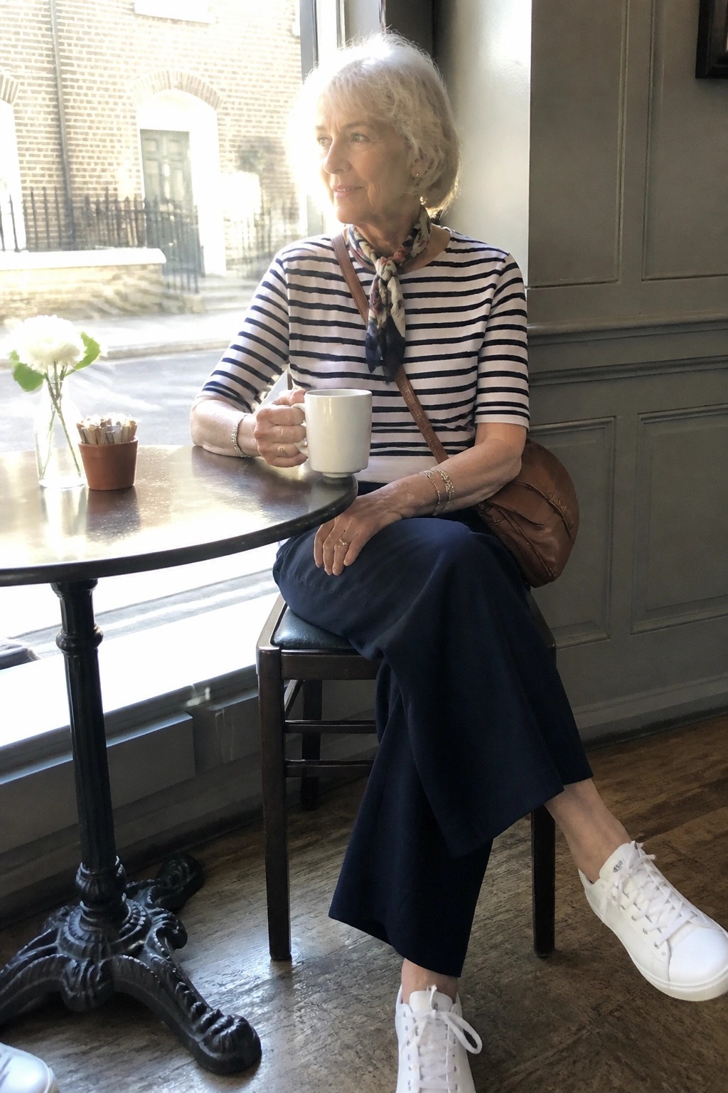 Woman over 50 in striped short sleeve top, navy wide leg pants, white sneakers, brown crossbody bag and patterned neck scarf sitting at a cafe table holding a mug.