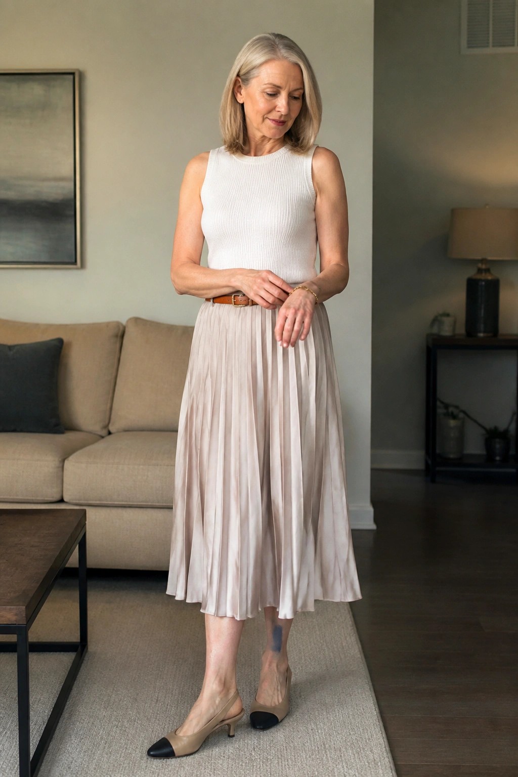 Gray-haired woman in white top and beige pleated skirt adjusts brown belt indoors