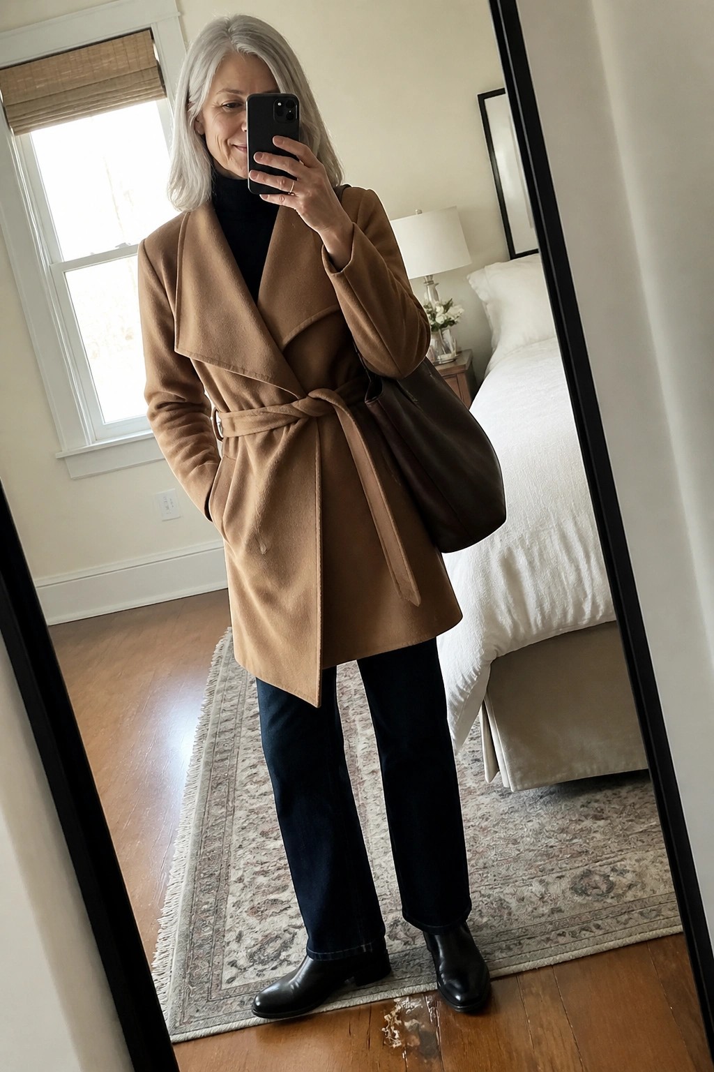 Woman with gray hair in tan belted coat taking mirror selfie in bedroom