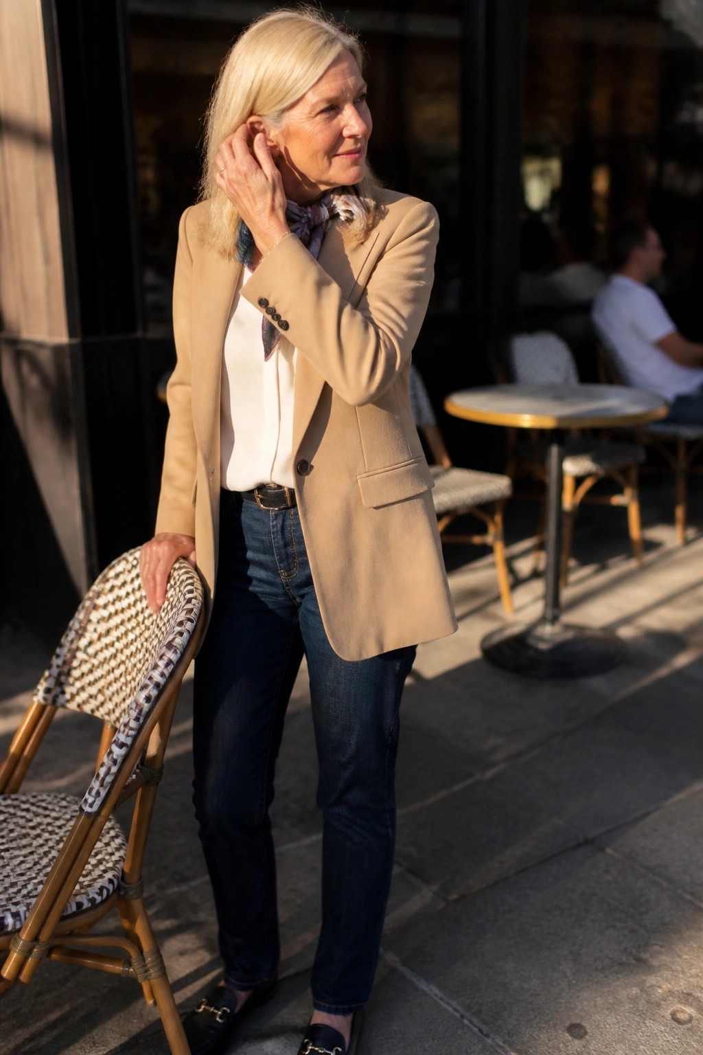 Stylish blonde woman in beige blazer and jeans adjusting earring by cafe chair.