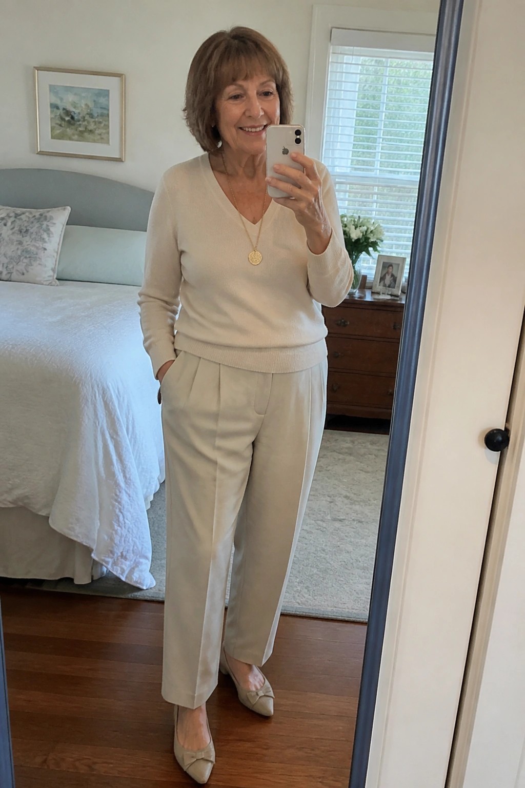 Smiling woman in cream sweater and pleated pants taking bedroom mirror selfie