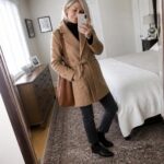 Woman with gray hair in tan belted coat taking mirror selfie in bedroom