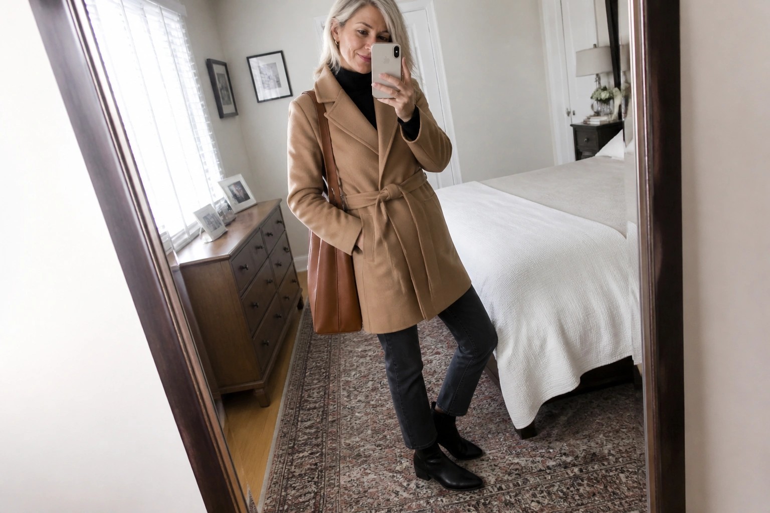 Woman with gray hair in tan belted coat taking mirror selfie in bedroom