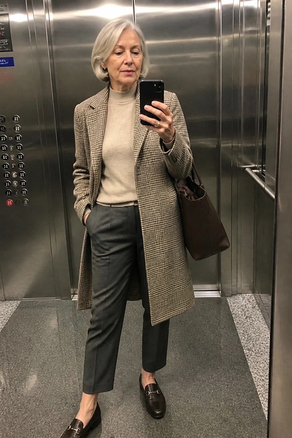 Gray-haired woman in houndstooth coat taking selfie in elevator with brown bag