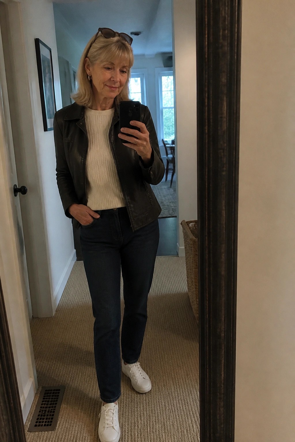 Woman in black leather jacket and white sweater taking mirror selfie.