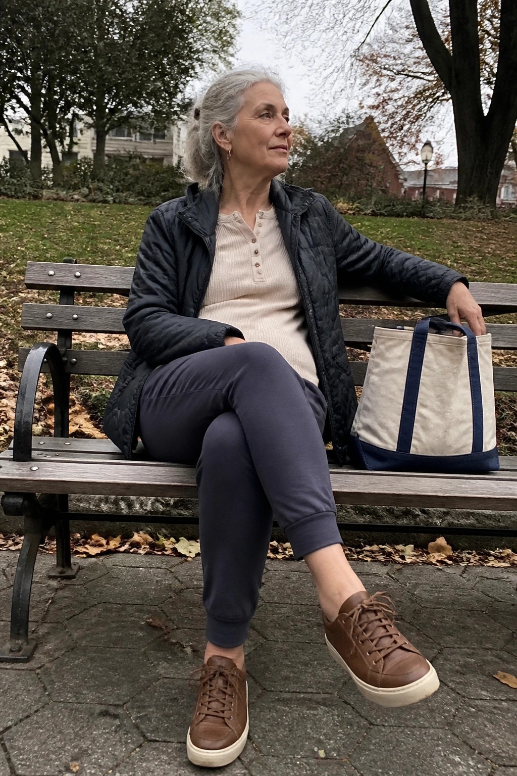 Gray-haired woman in quilted jacket sits on park bench beside canvas tote bag.