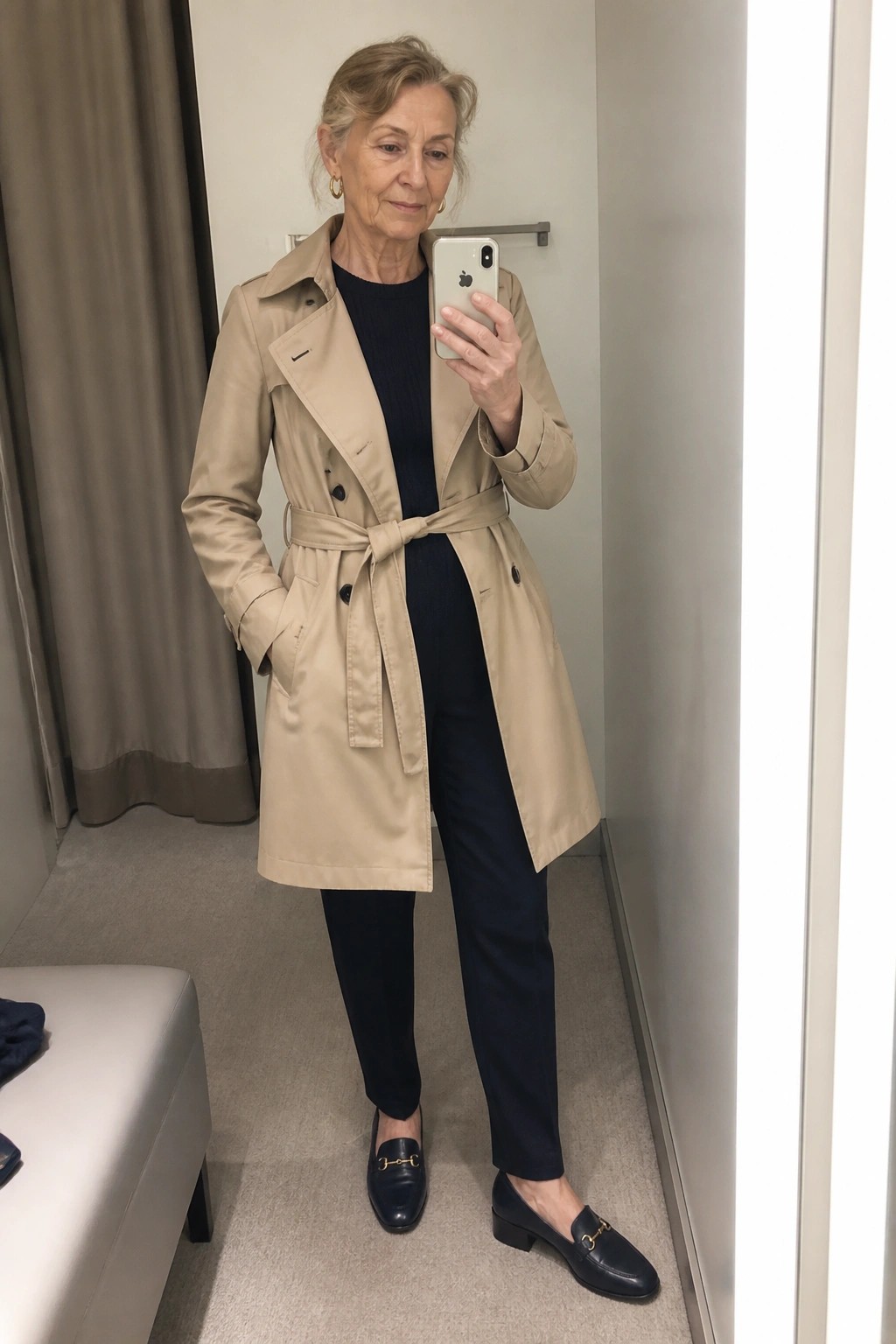 Woman in beige trench coat taking iPhone mirror selfie in fitting room