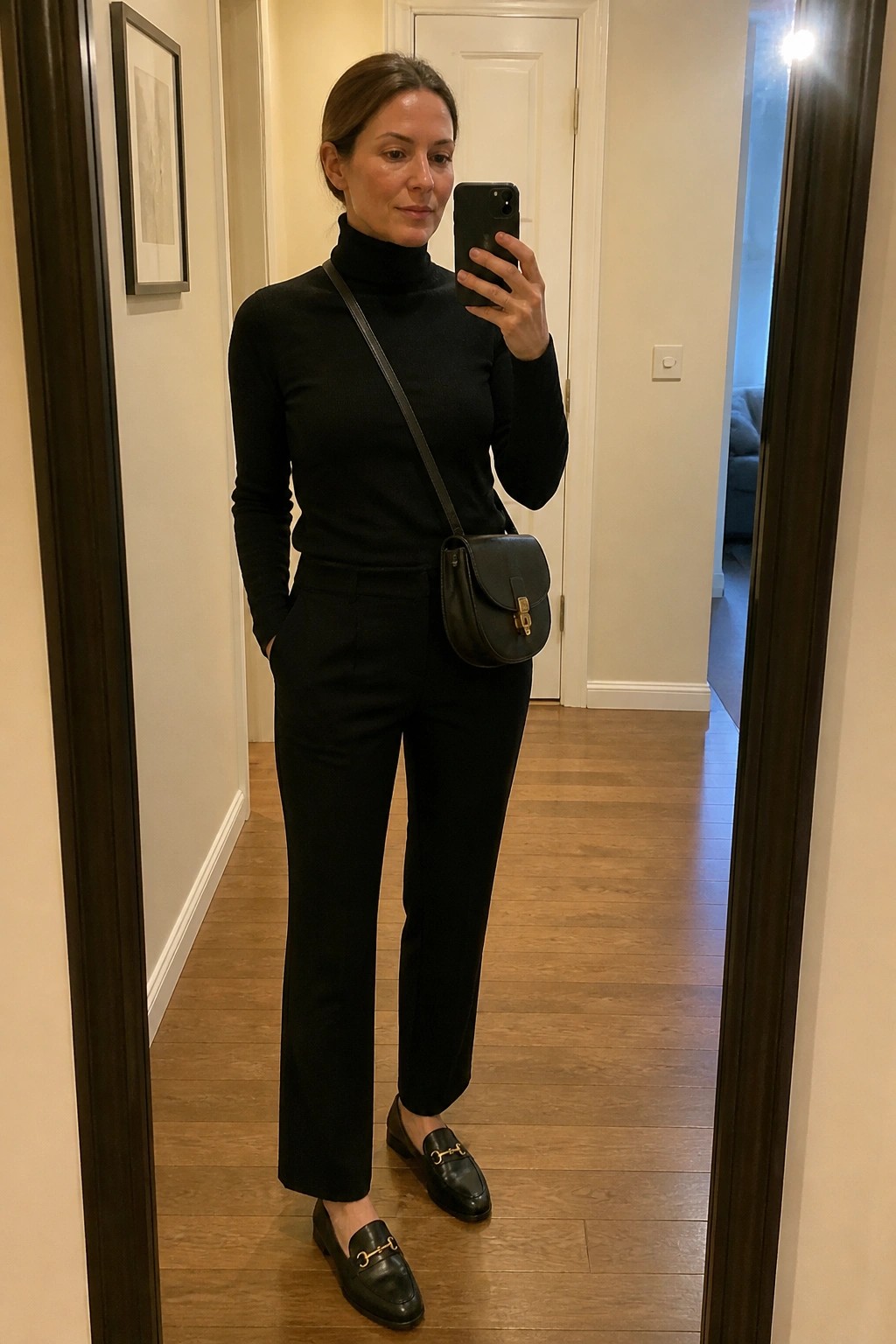 Woman in black turtleneck, pants, and loafers taking mirror selfie with crossbody bag