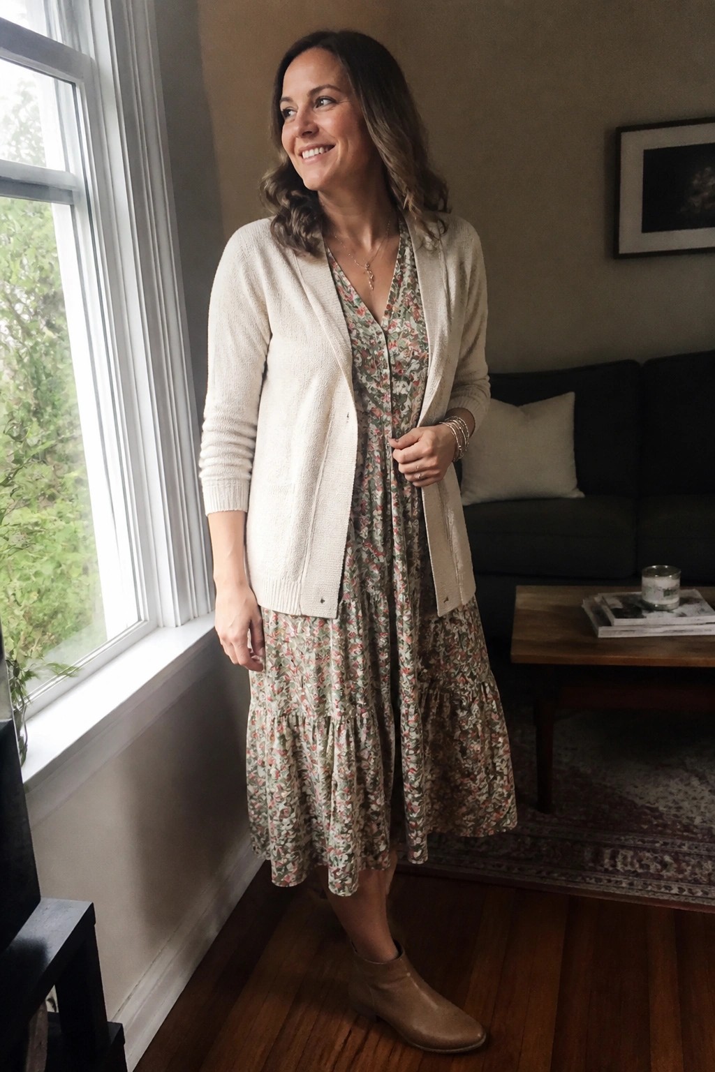 Smiling woman in floral dress and cream cardigan standing by window in living room