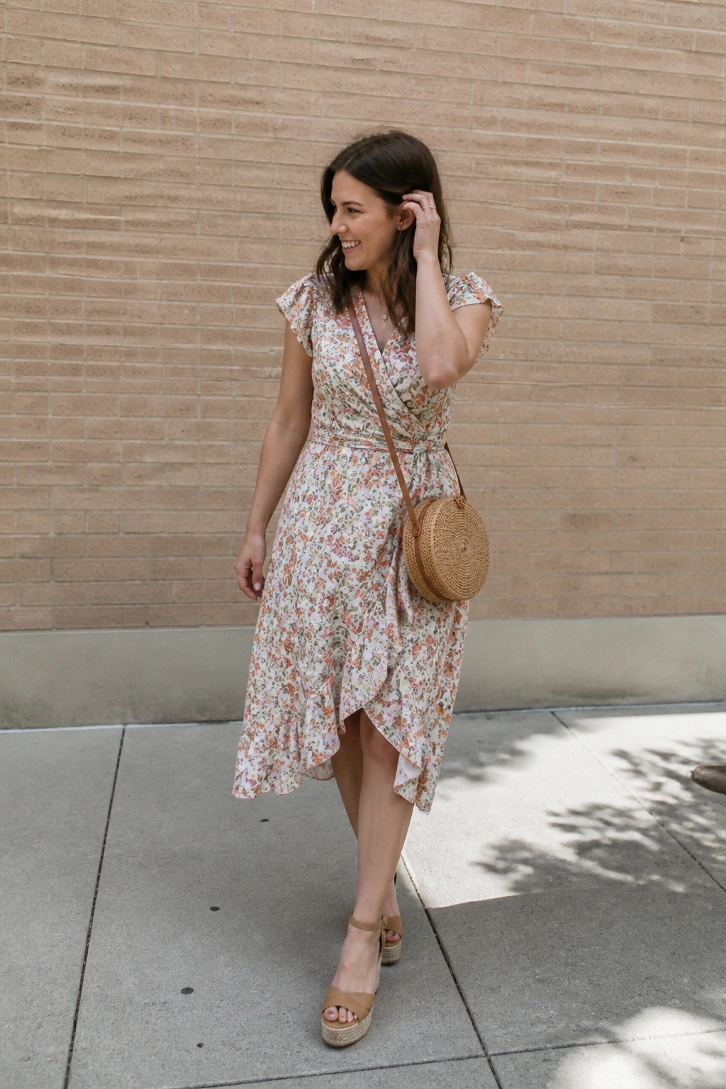 Woman in a floral wrap dress featuring ruffled cap sleeves and an asymmetrical hem paired with a round straw crossbody bag and beige wedge sandals.