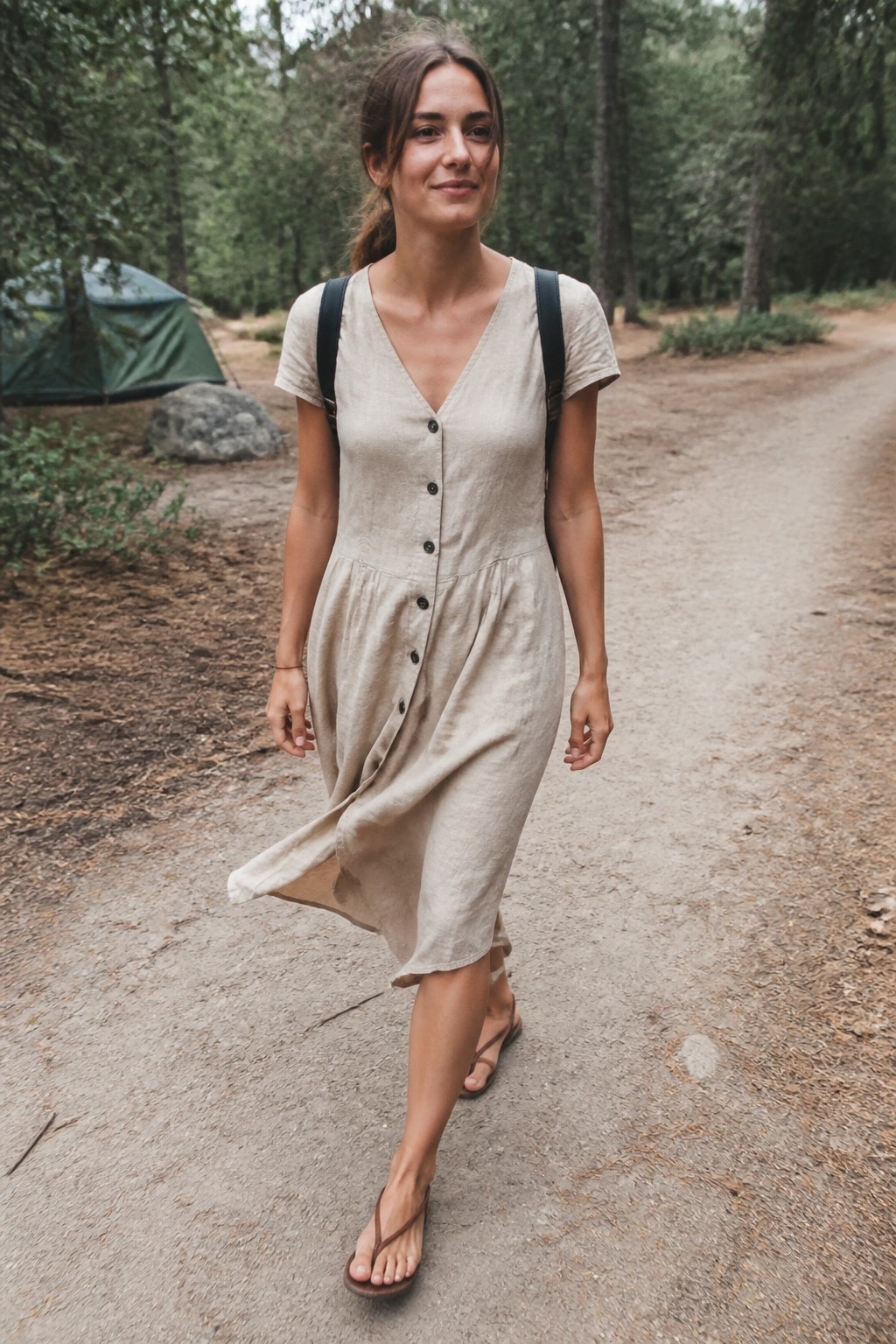 Woman in beige buttoned dress walking on forest dirt path with backpack