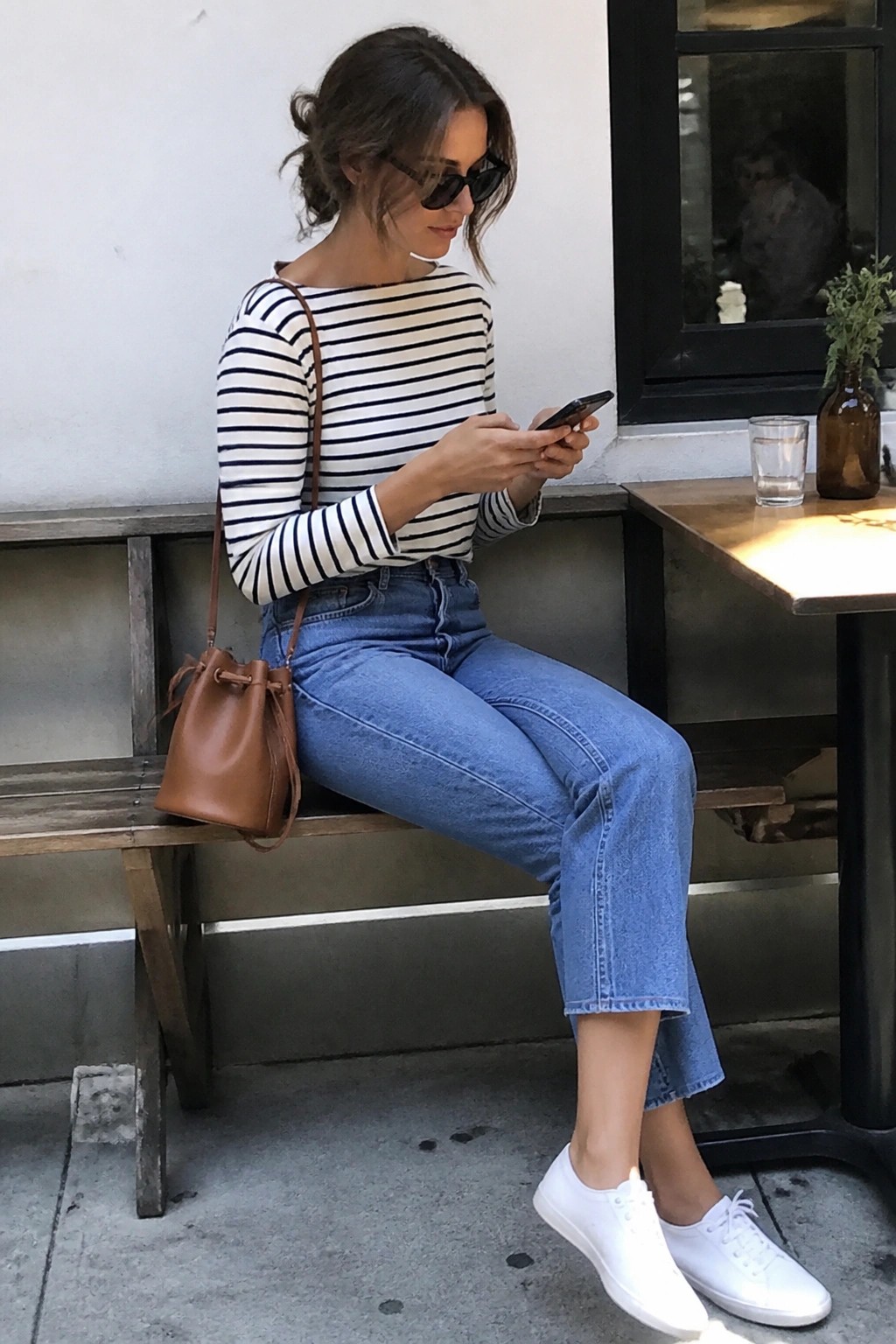 Woman in striped shirt and jeans sits on bench checking phone with brown bag