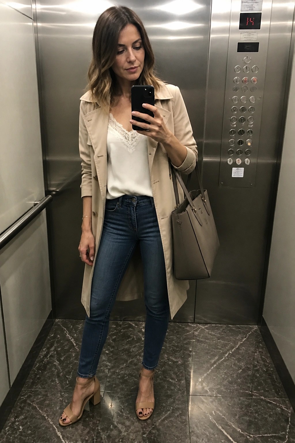 Woman in beige trench coat taking selfie in elevator with gray handbag