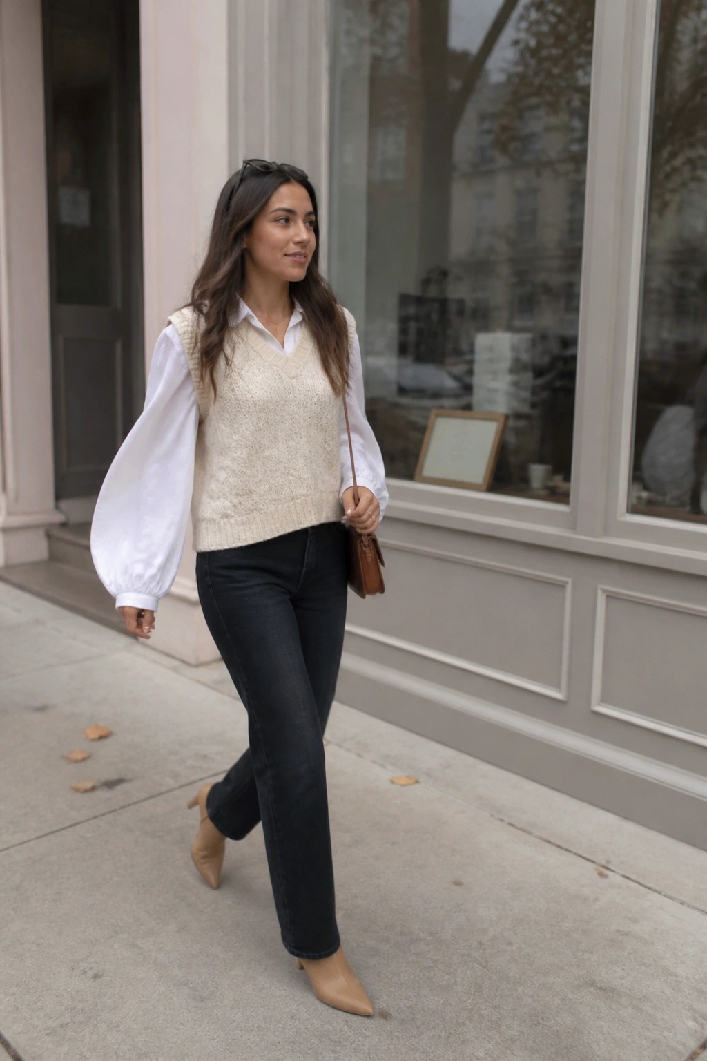 Woman in white blouse, cream knit vest, dark jeans, and beige boots on city sidewalk