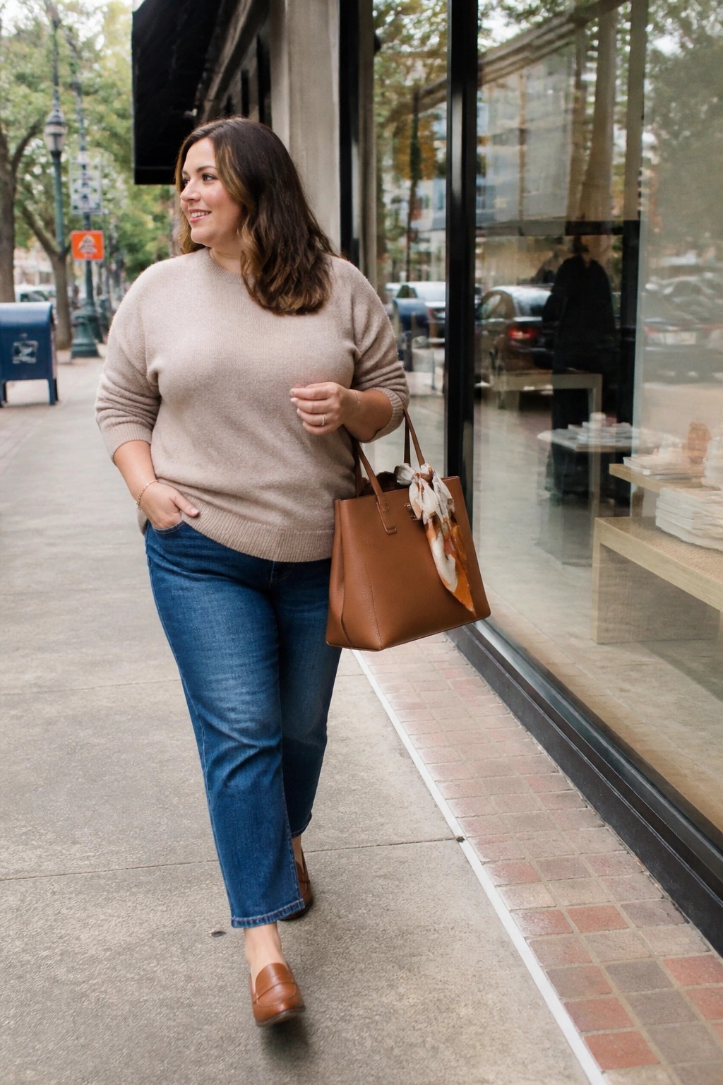 Woman in beige sweater and blue jeans carrying brown handbag on city sidewalk