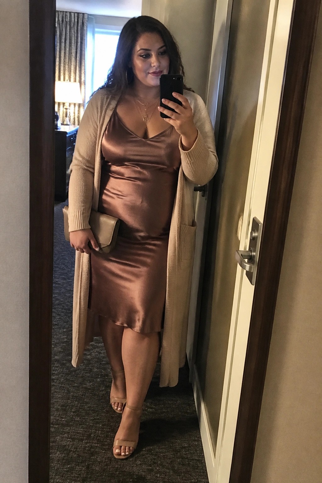 Woman in bronze satin dress and beige cardigan taking mirror selfie with phone