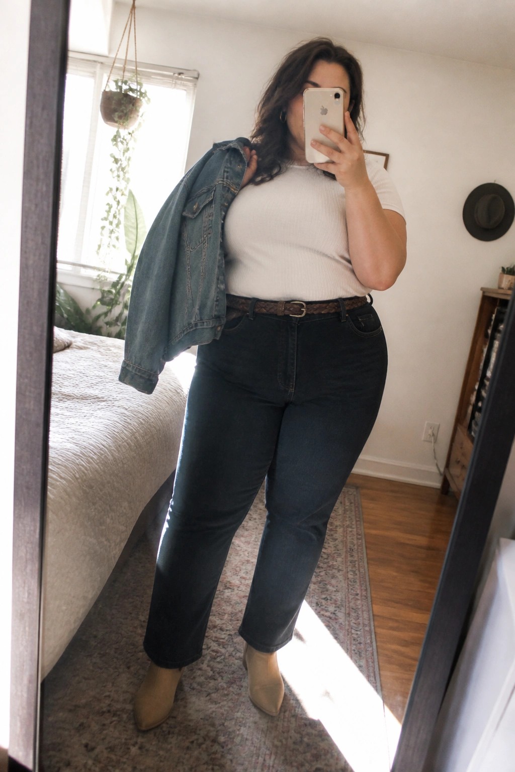A plus size woman in a mirror selfie wearing a white ribbed top, dark blue jeans with a brown belt, holding a denim jacket over one shoulder, and beige ankle boots.