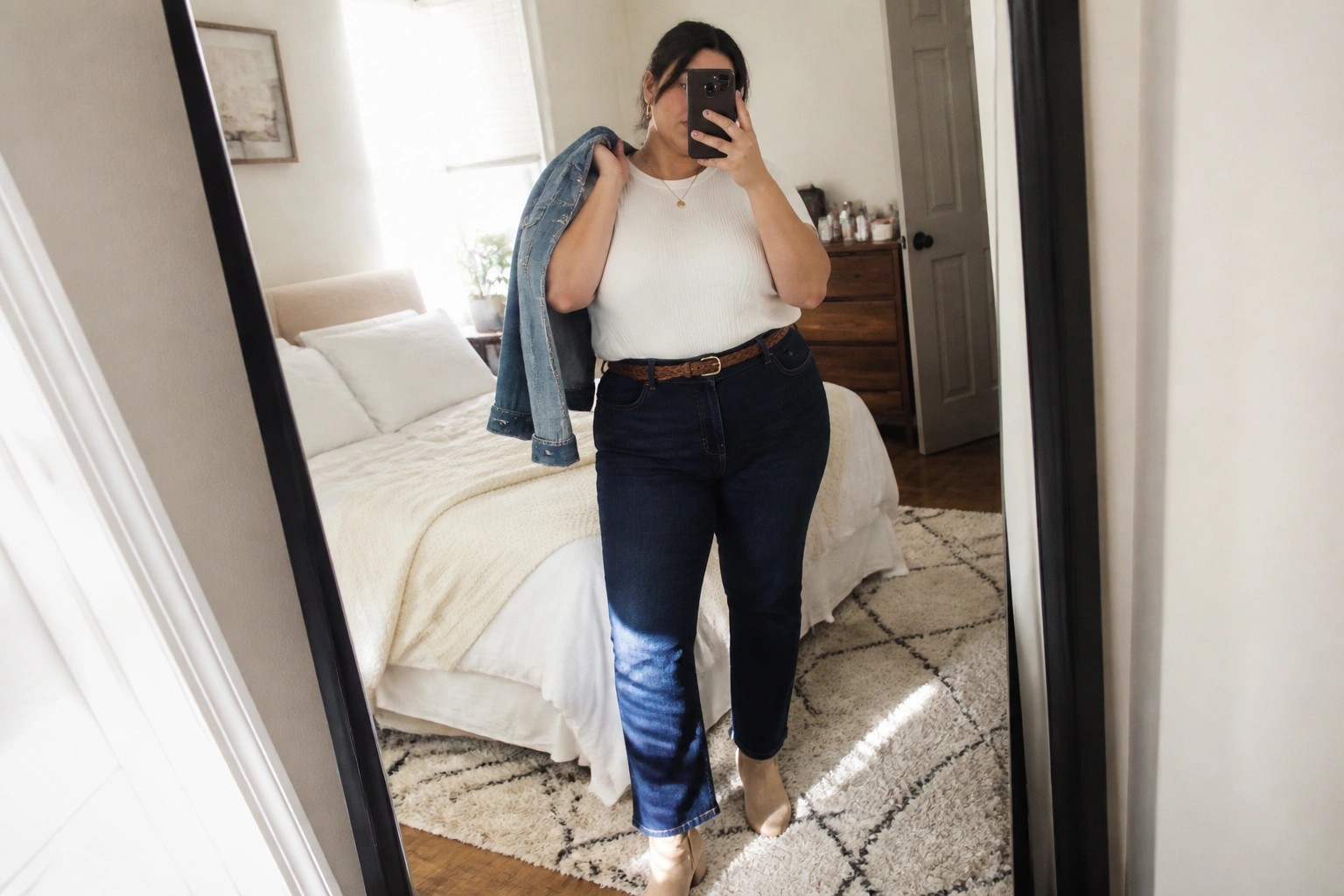 A plus size woman in a mirror selfie wearing a white ribbed top, dark blue jeans with a brown belt, holding a denim jacket over one shoulder, and beige ankle boots.