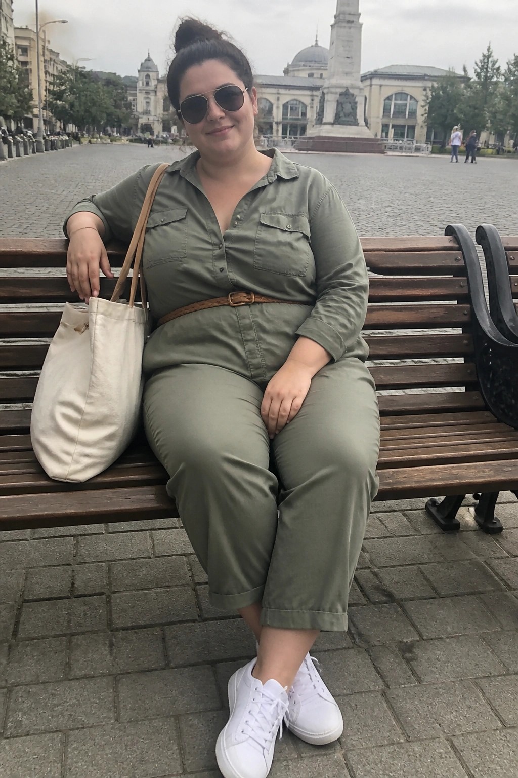 Woman in green jumpsuit and sunglasses with tote bag on city bench
