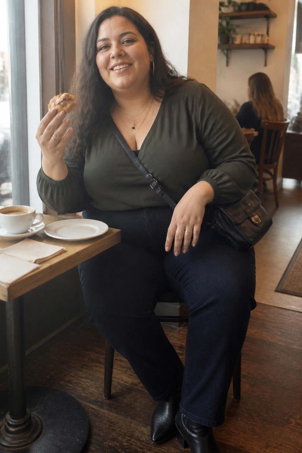 Smiling woman in green top holds pastry at cafe table with coffee cup