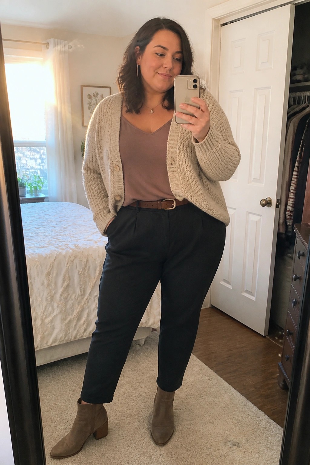 Woman in beige cardigan and black pants taking bedroom mirror selfie with phone.