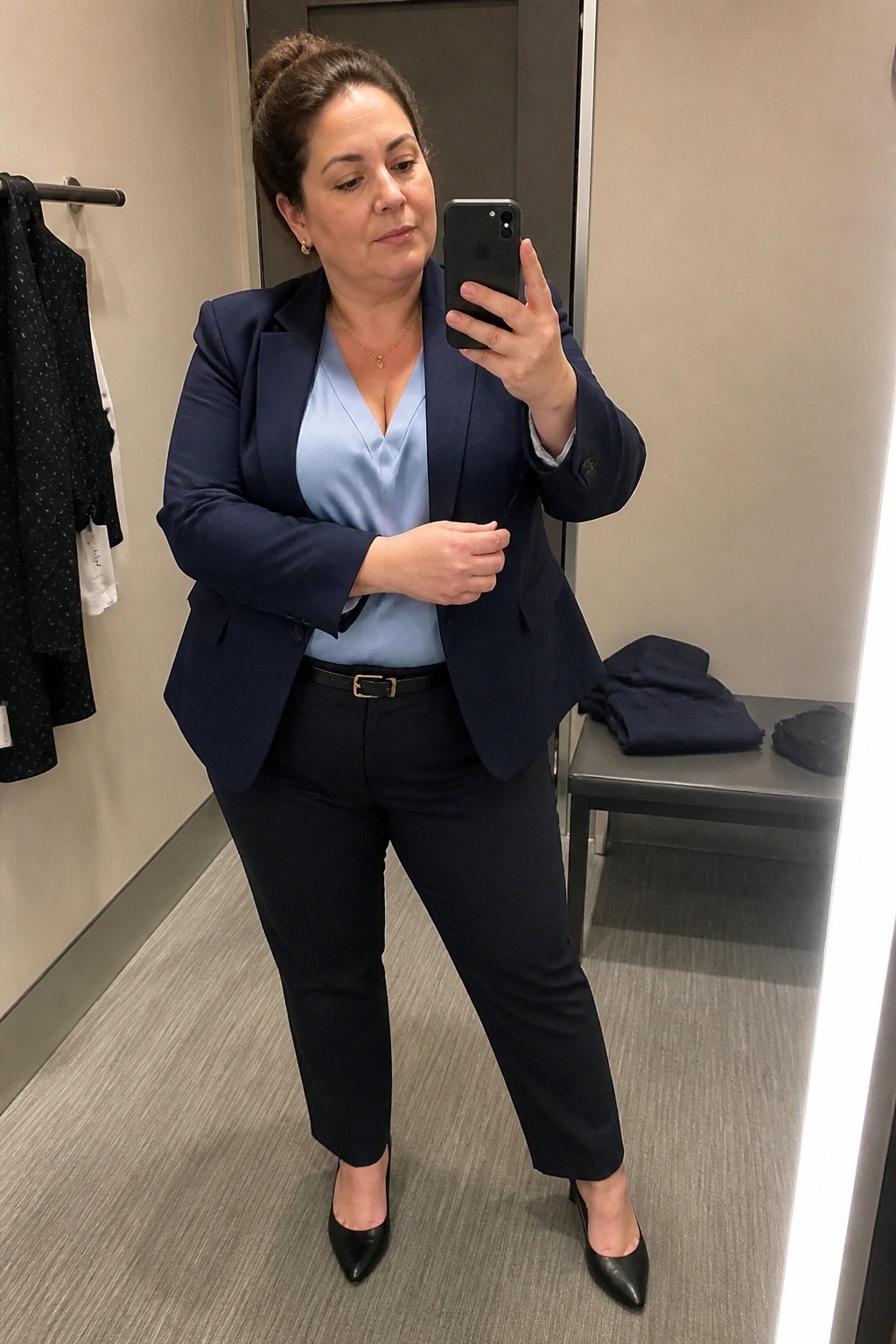 Woman wearing a navy blazer over a light blue satin top with matching navy pants, black belt, and black pointed heels while taking a mirror selfie in a fitting room.