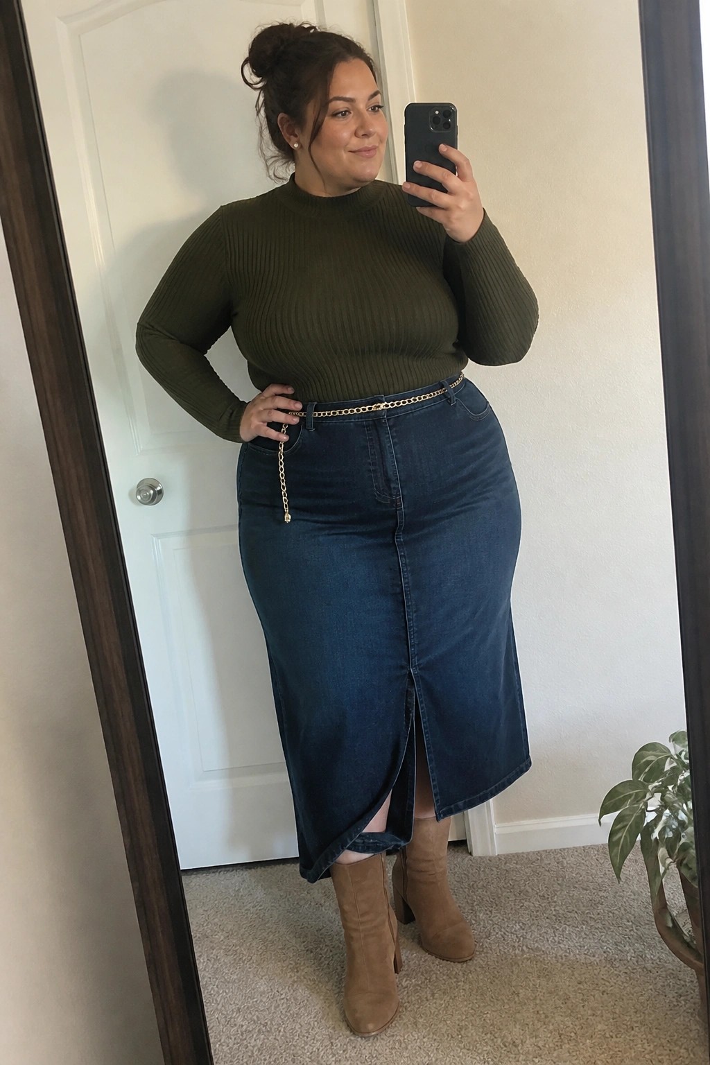 A plus size woman wearing an olive green ribbed sweater, blue denim midi skirt with gold chain belt, and tan suede ankle boots, taking a mirror selfie.