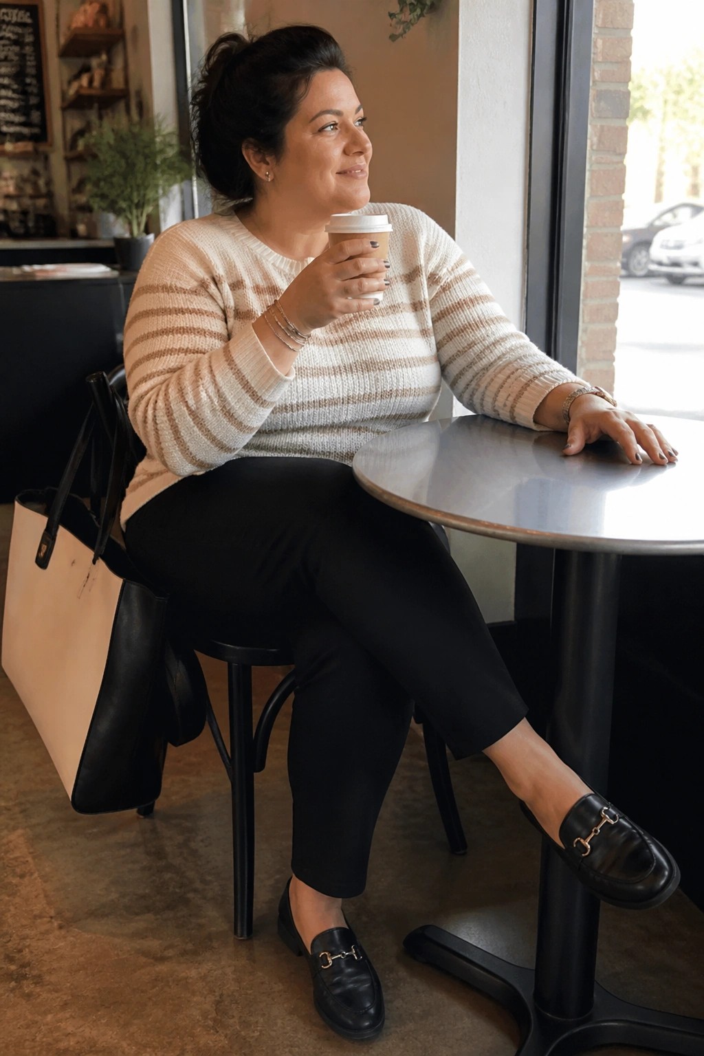 Woman in striped sweater holding coffee at round cafe table with tote bag