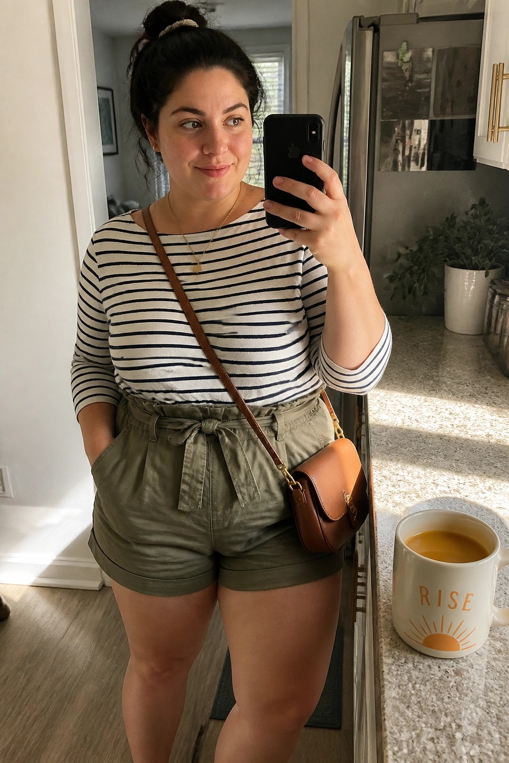 Woman in striped top and green shorts taking mirror selfie in kitchen with rise mug