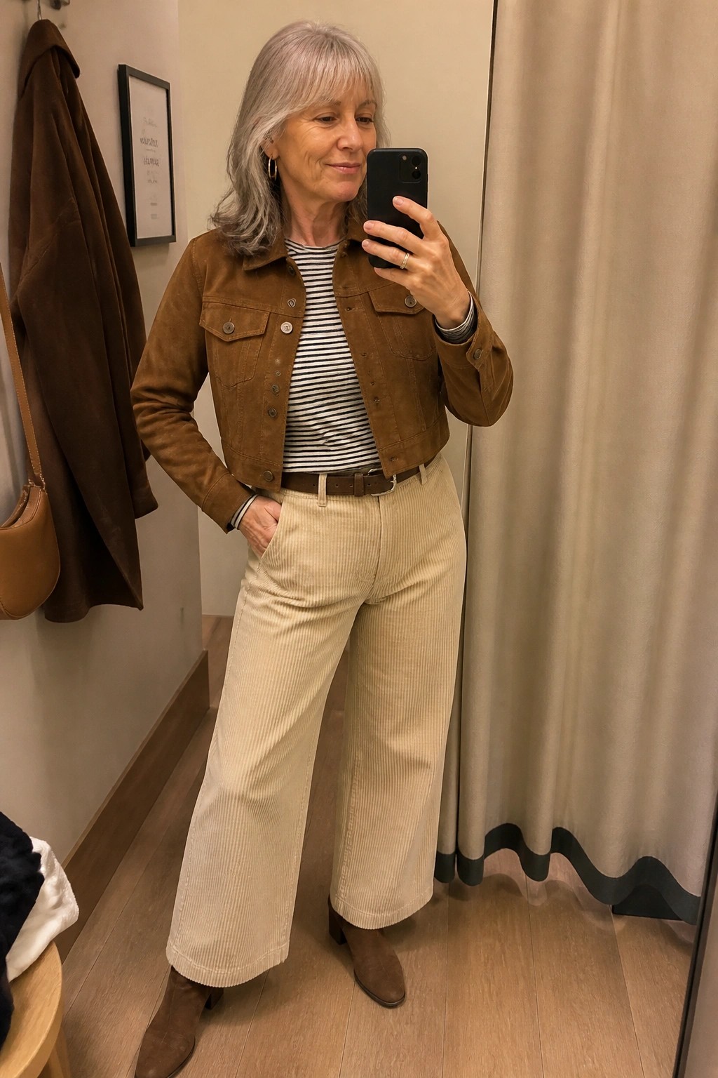 Gray-haired woman in brown suede jacket and beige corduroy pants taking mirror selfie