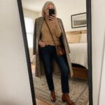 Silver-haired woman in plaid coat and tan turtleneck taking bedroom mirror selfie