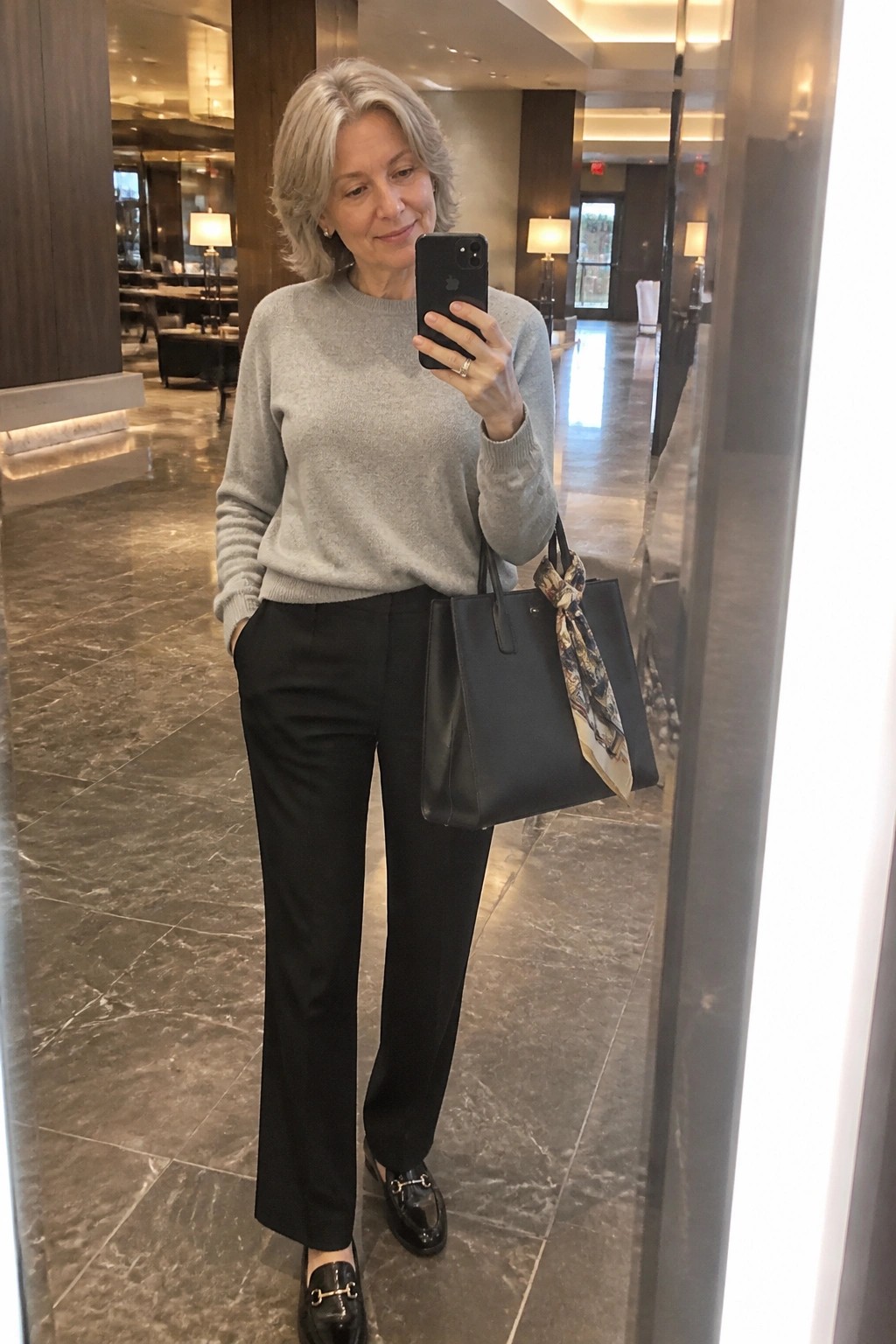 Woman over 60 wearing a light gray knit sweater, black straight-leg pants, black loafers with gold hardware, and carrying a black tote bag with a patterned scarf tied on the handle.