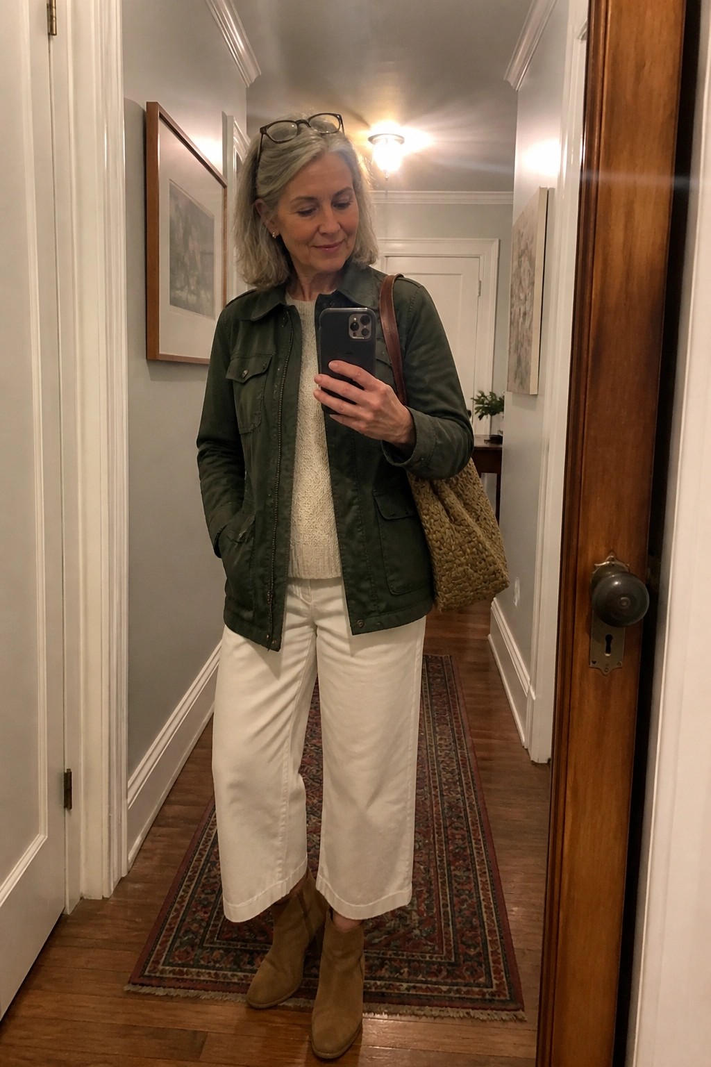 Gray-haired woman in green jacket and white pants taking mirror selfie in hallway