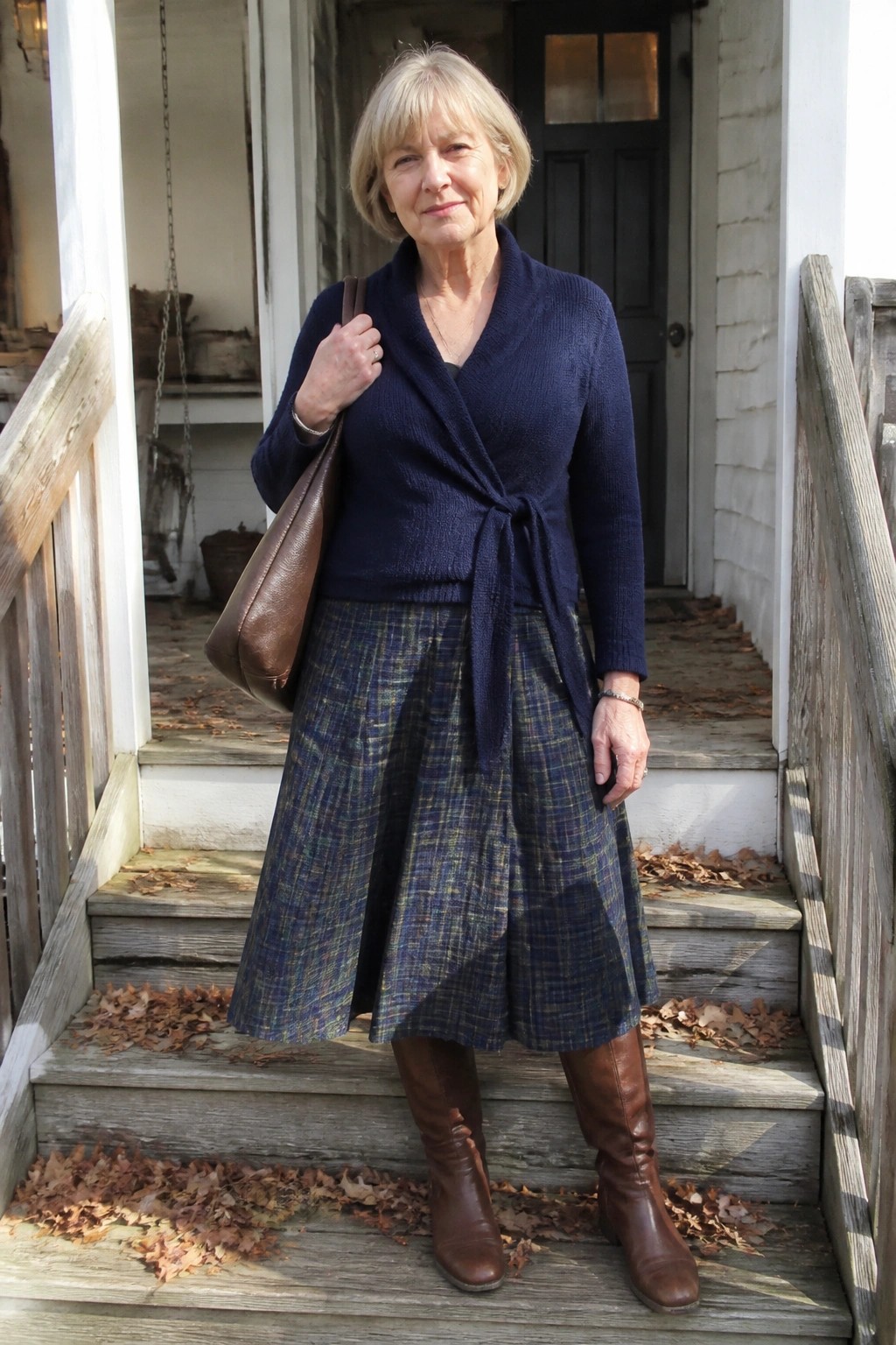 Elderly woman in navy wrap sweater and plaid skirt on leaf-covered porch steps.
