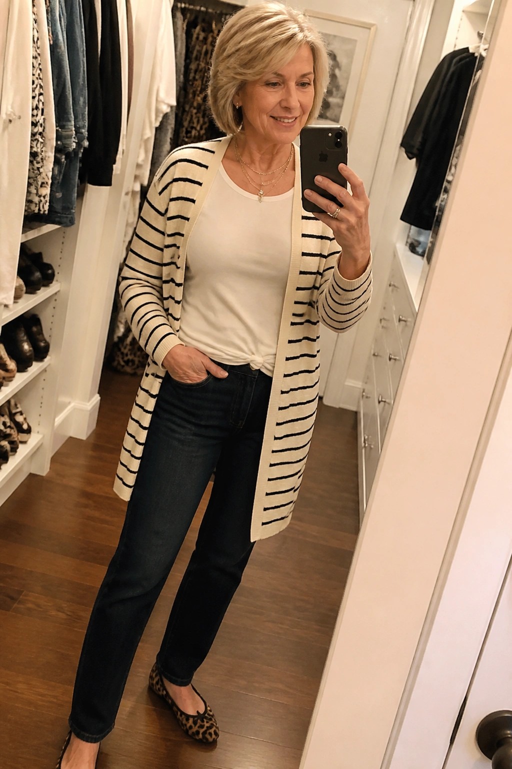 Woman over 60 wearing a long cream cardigan with black horizontal stripes over a white knotted tee, dark blue jeans, leopard print flats, and layered gold necklaces while taking a mirror selfie in a closet.
