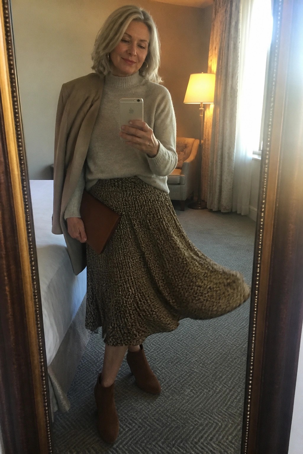 Woman over 60 wearing a beige turtleneck sweater under a draped taupe blazer paired with a brown patterned midi skirt brown suede ankle boots and a matching clutch while taking a mirror selfie.