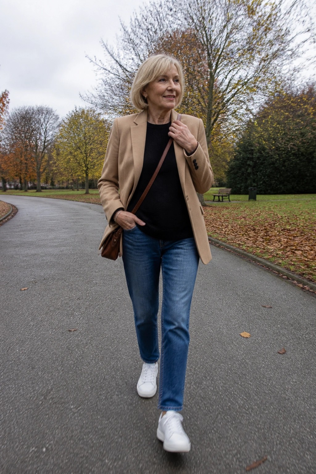 Blonde woman in tan blazer, black top, blue jeans, and white sneakers in park