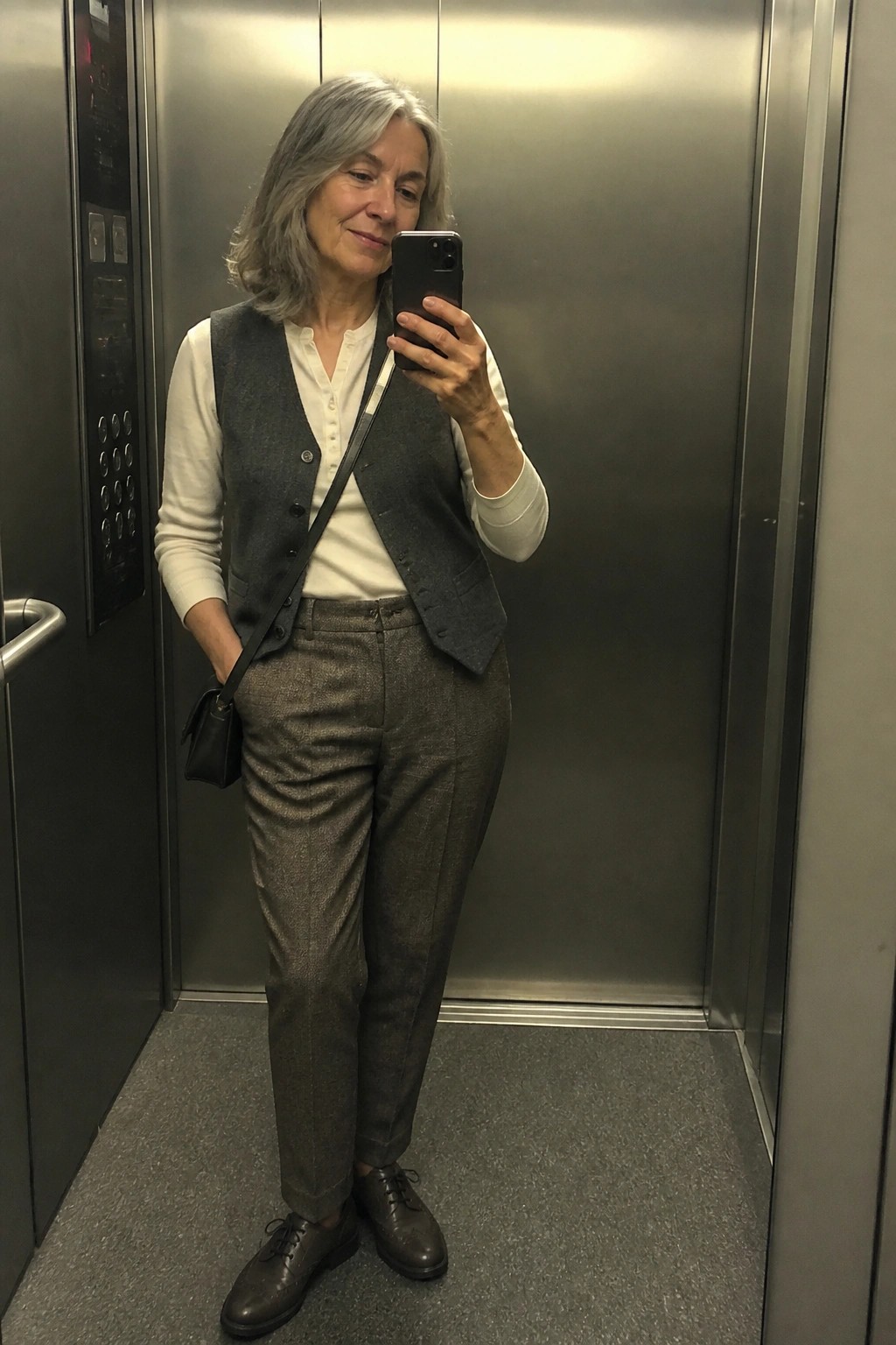 Gray-haired woman in gray vest taking phone selfie inside elevator