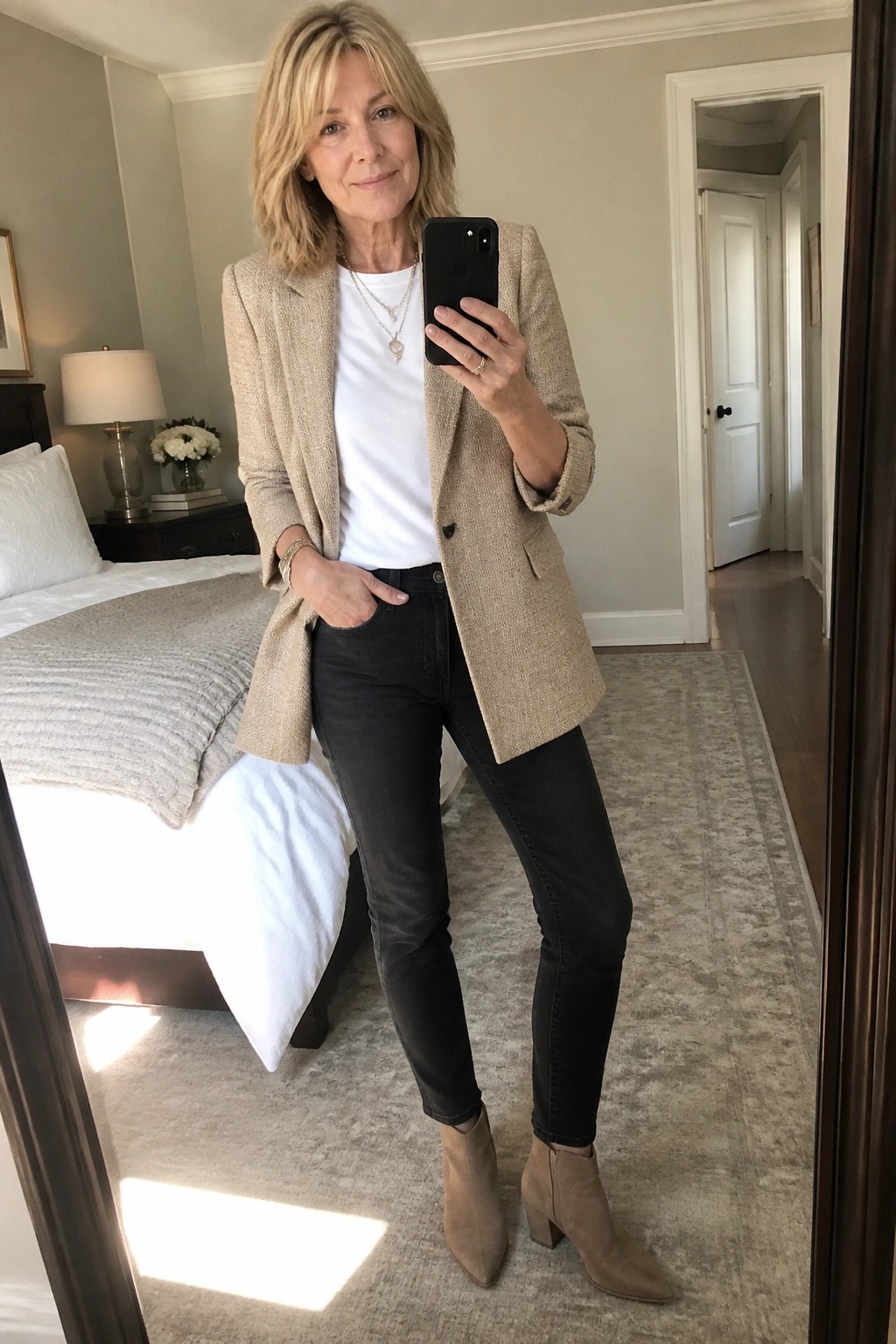 Blonde woman in beige blazer, white tee, and black jeans taking bedroom mirror selfie
