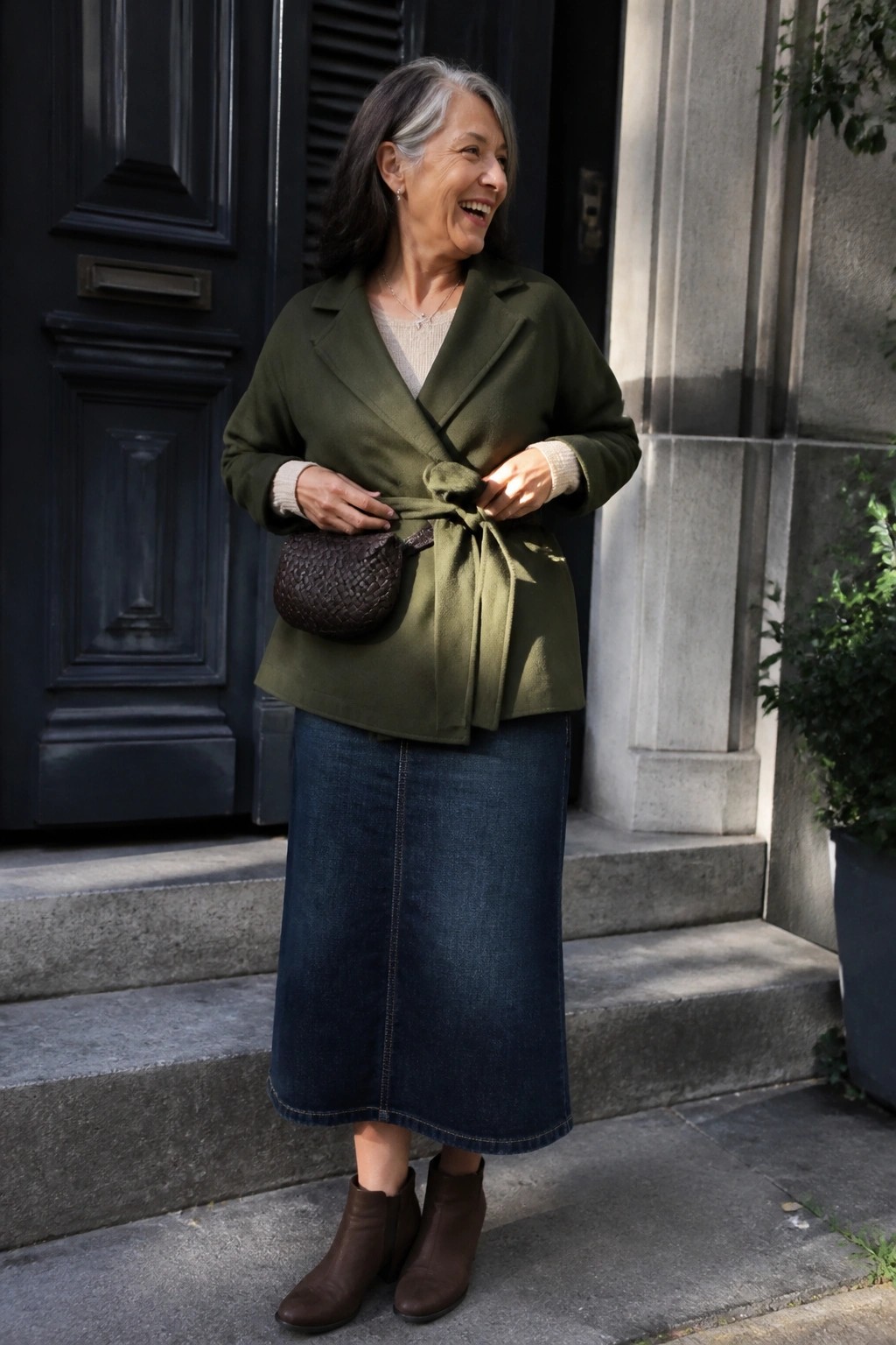 Woman over 60 wearing an olive green wrap jacket tied at the waist over a cream sweater and dark blue denim midi skirt with brown ankle boots and a woven brown crossbody bag.
