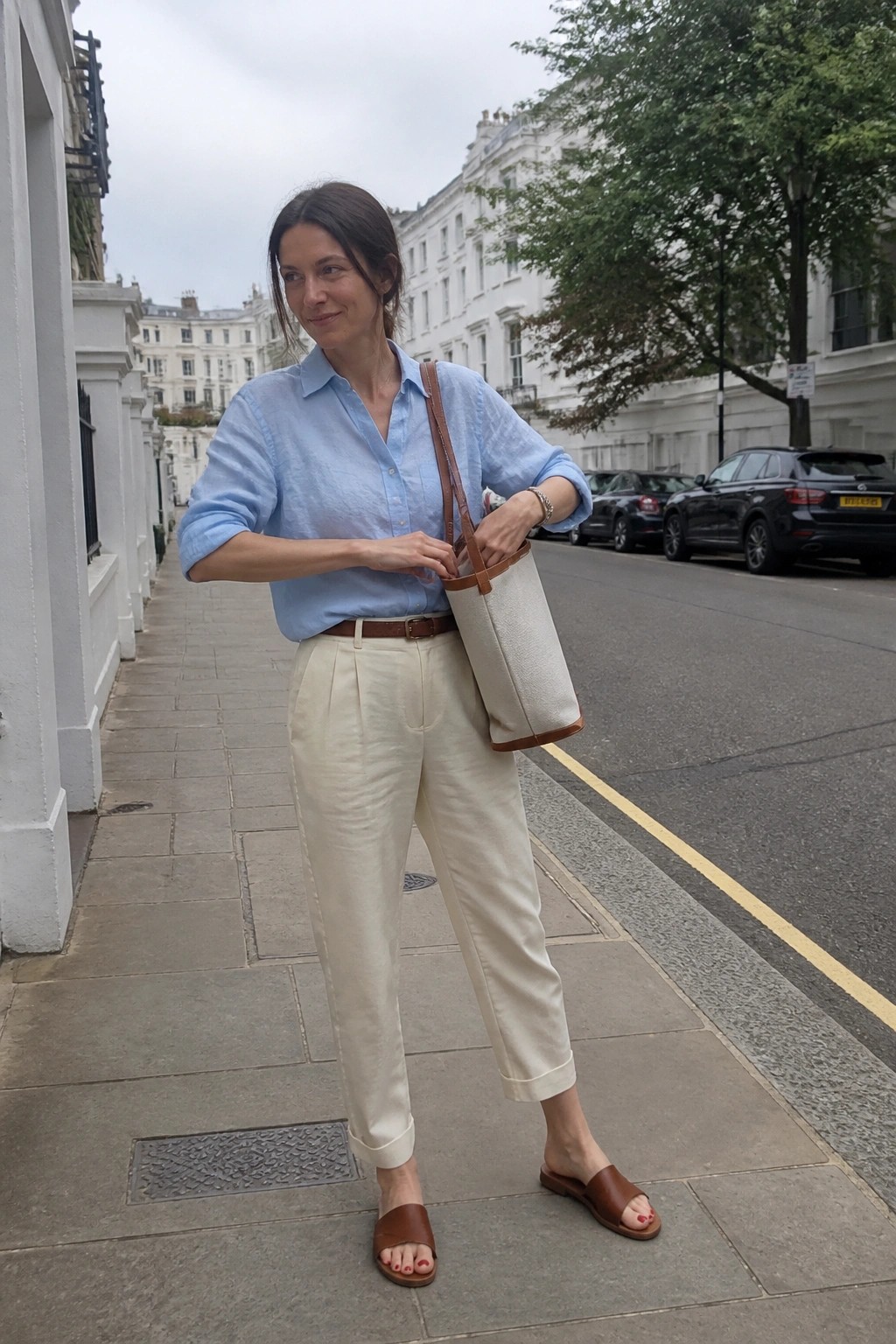 Woman wearing light blue button down shirt tucked into cream pleated trousers with brown leather belt, matching brown slides, and carrying a beige canvas tote bag on a city sidewalk.
