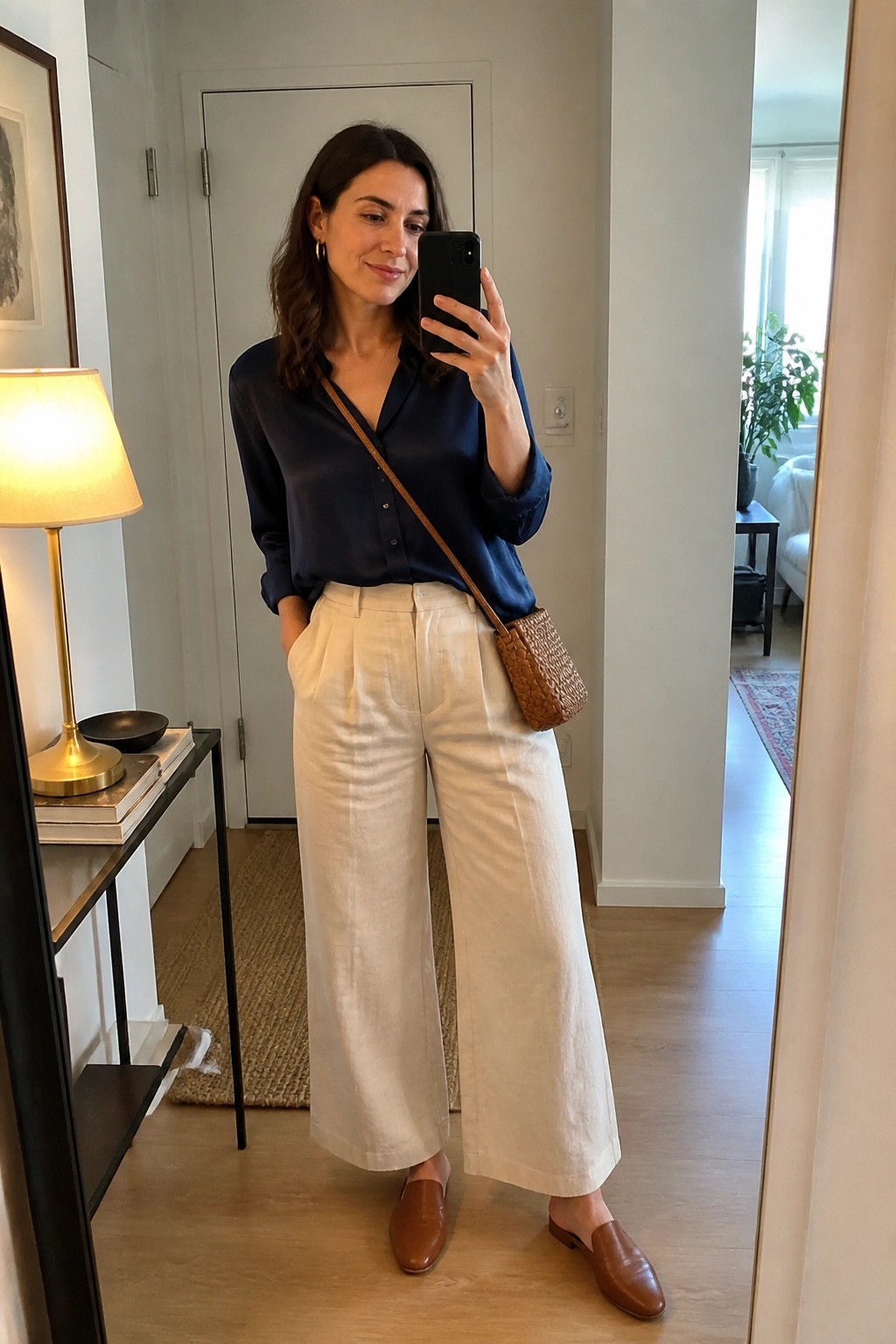 Woman in navy blue silk button-up shirt tucked into cream wide-leg linen pants with brown leather mules and woven brown crossbody bag taking a mirror selfie indoors.