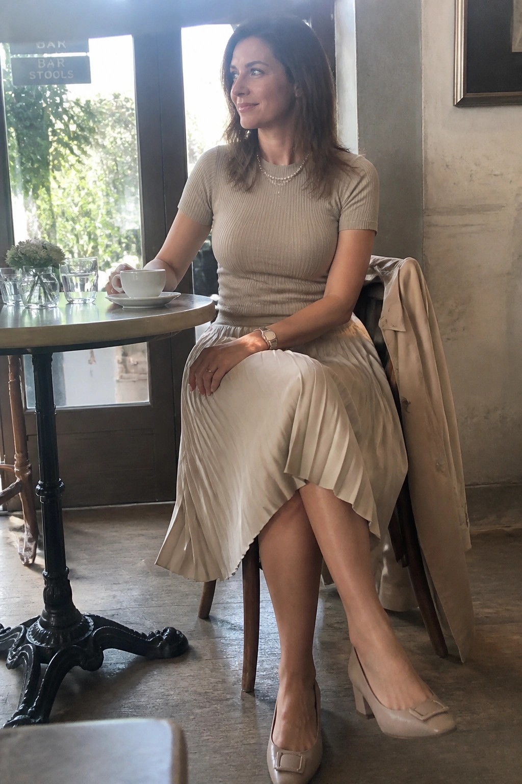 Woman in beige top and pleated skirt holds coffee at cafe table