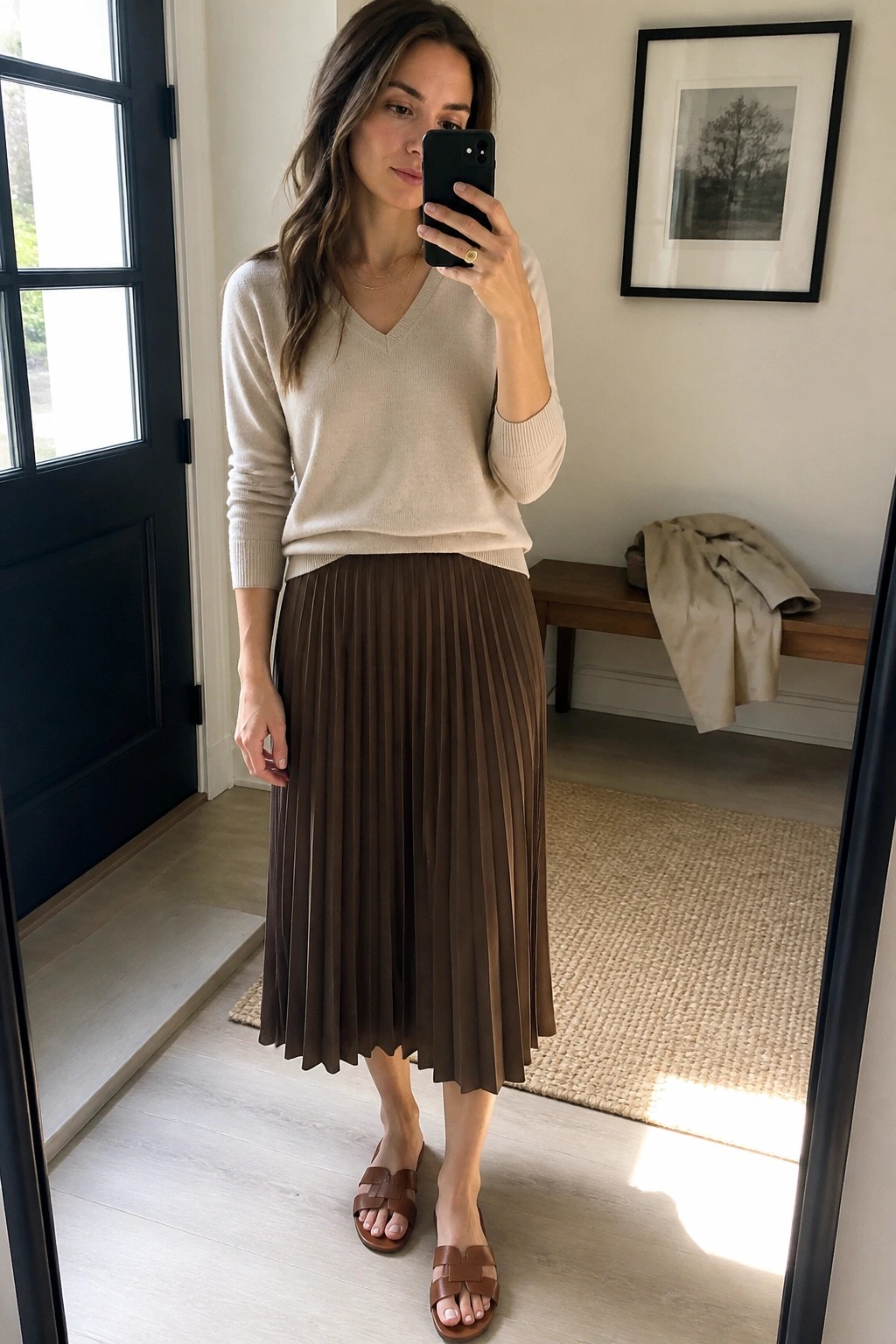 Woman in beige sweater and brown pleated skirt taking mirror selfie with phone