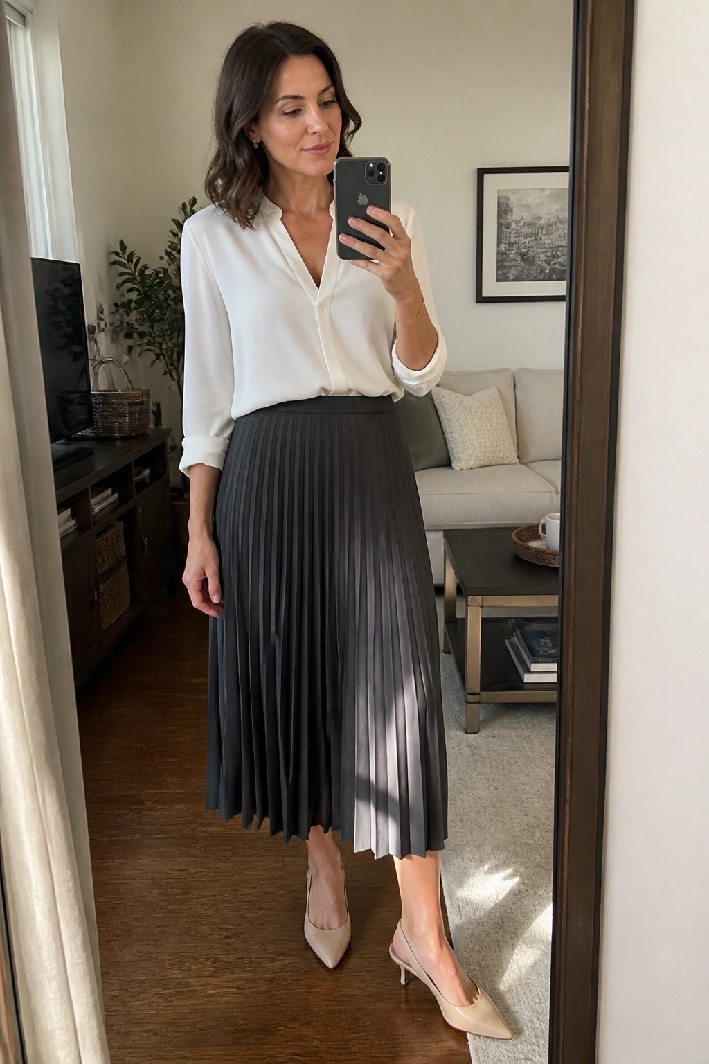 Woman in white blouse and gray pleated skirt taking mirror selfie in living room