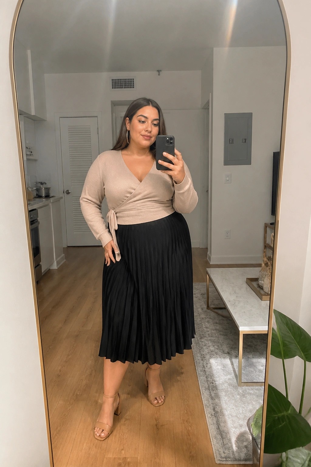 Woman in beige wrap sweater and black pleated skirt taking mirror selfie in apartment.