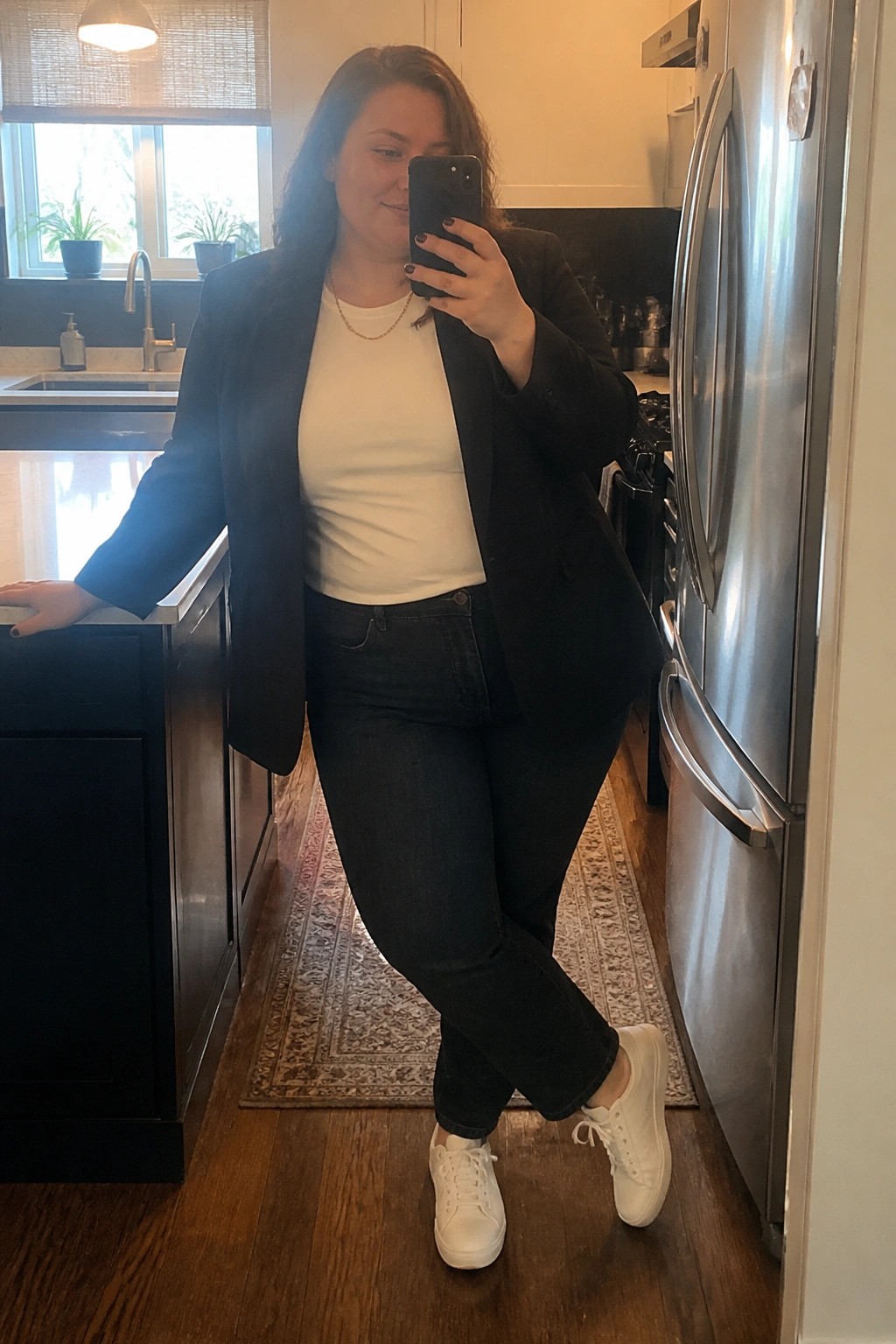 Woman in black blazer, white tee, and jeans taking kitchen mirror selfie