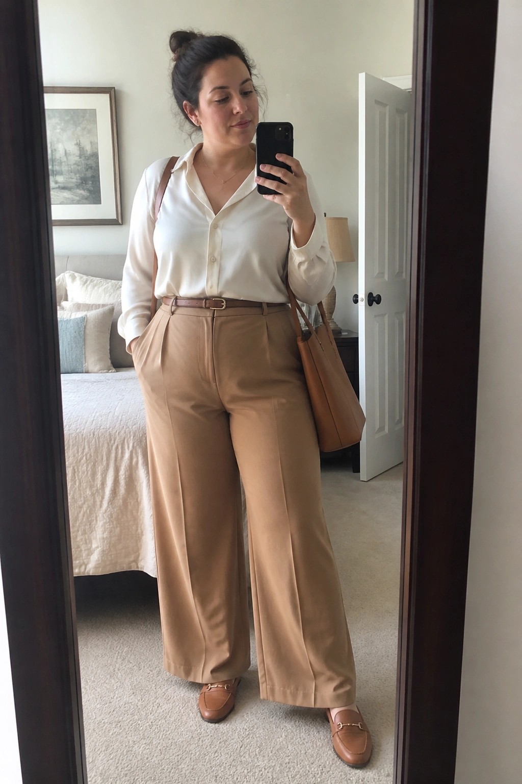 Plus size woman in mirror selfie wearing an ivory button down shirt tucked into high waisted wide leg tan trousers with a brown leather belt, matching loafers, and a brown tote bag.