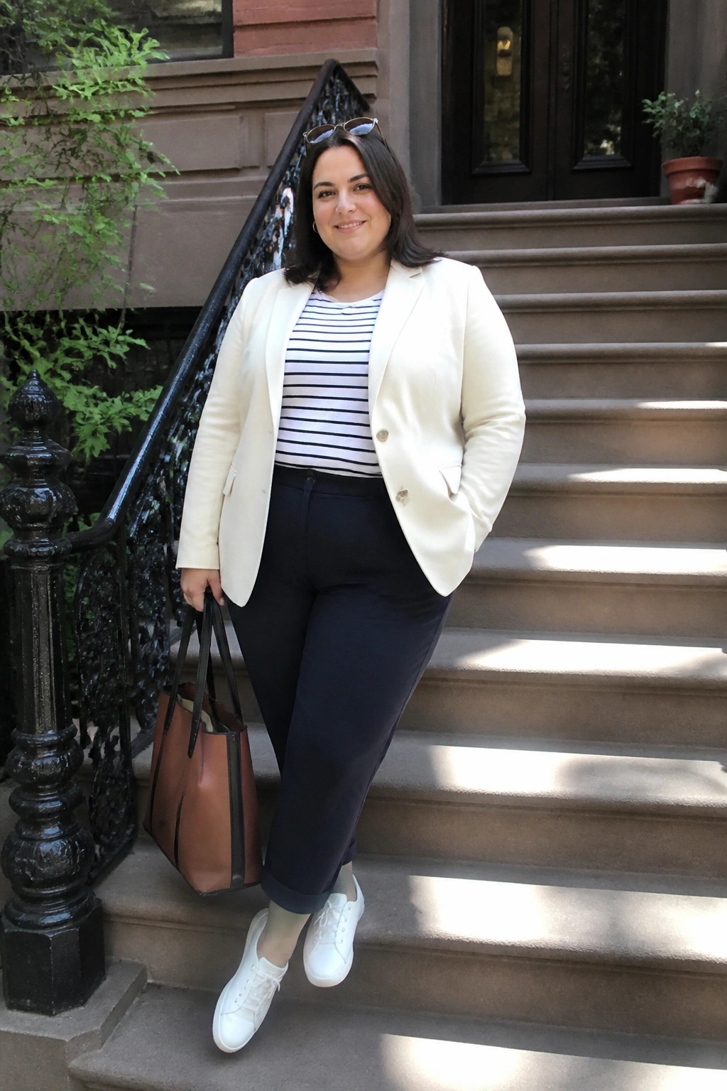 Plus size woman in cream blazer layered over black and white striped top with navy cuffed pants white sneakers and brown leather tote bag standing on stone steps.